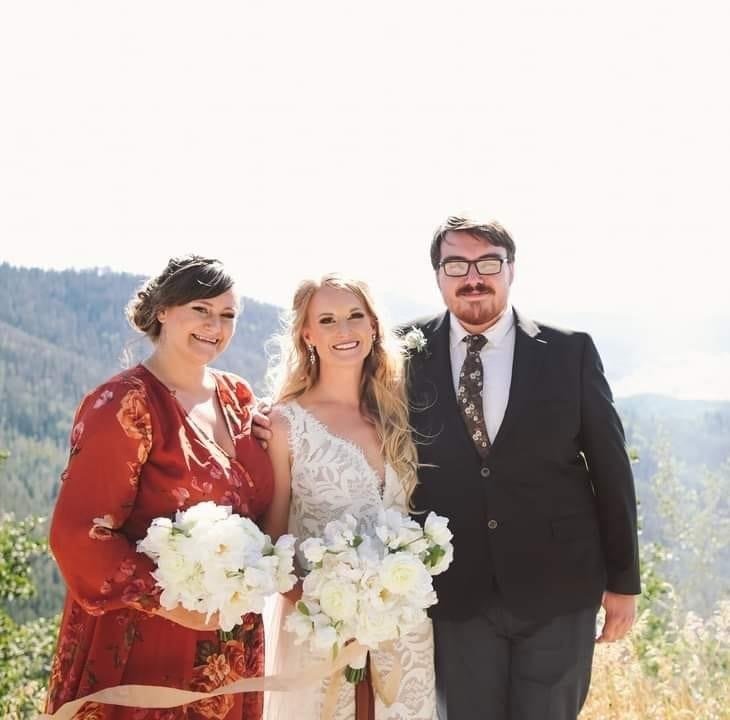 Three people standing outdoors with a scenic mountainous background, dressed in formal attire for a wedding, holding bouquets of white flowers.