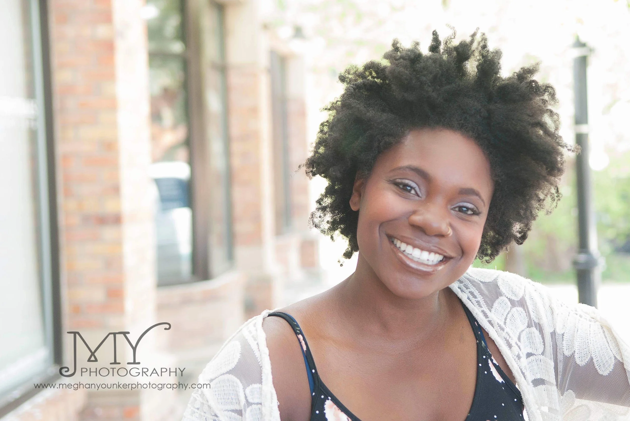 A smiling woman with natural curly hair and a nose ring, wearing a white lace cardigan and a black floral top outdoors near brick buildings.