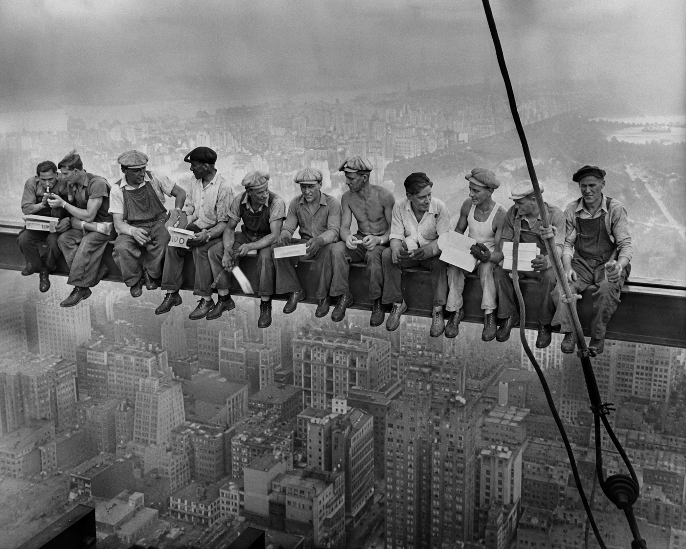Black and white photo of construction workers sitting on a steel beam high above a city, during the building of a skyscraper, with a cityscape in the background.