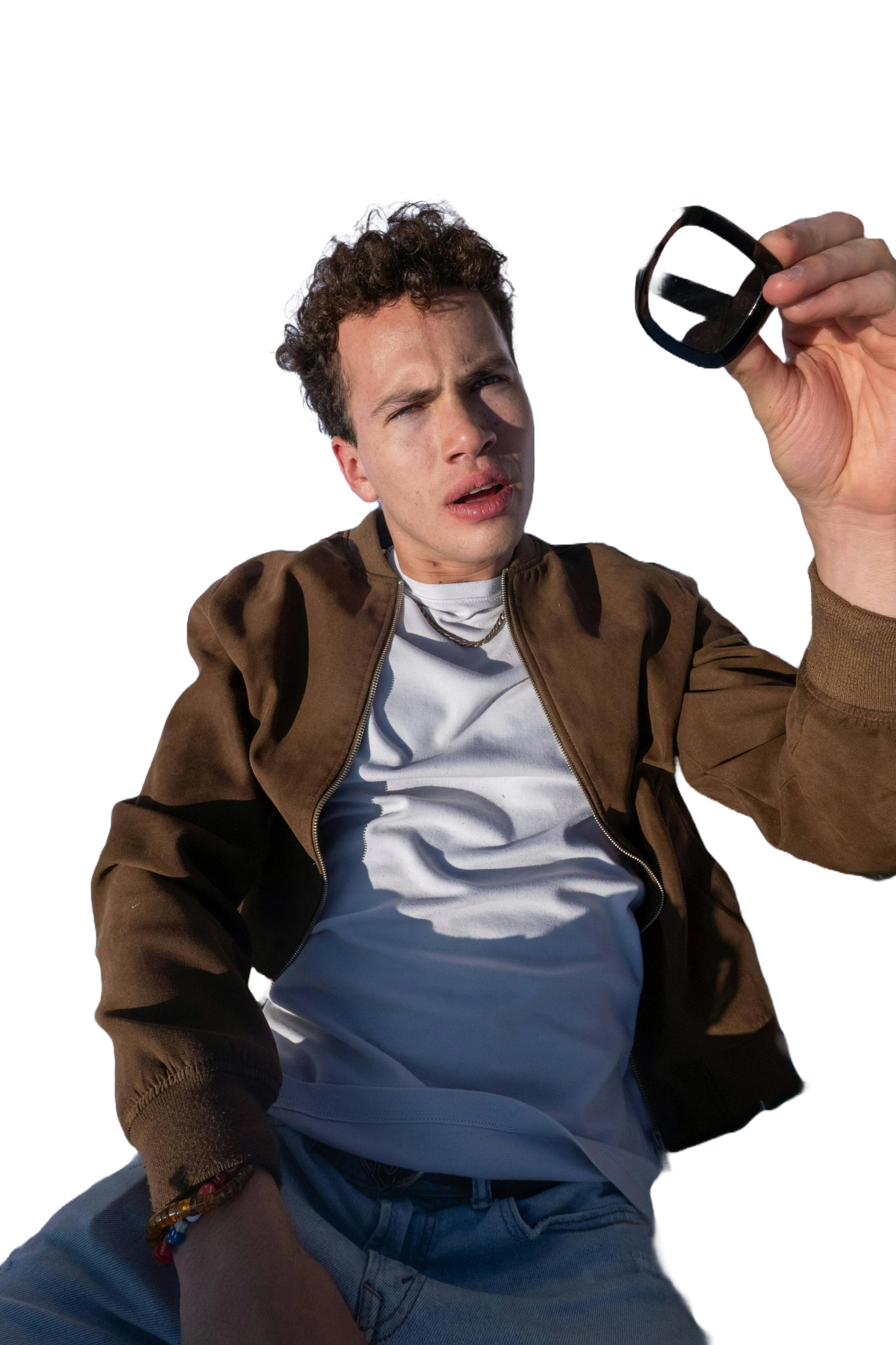 Young man with curly hair holding sunglasses, making a confused or puzzled expression while looking at the camera.