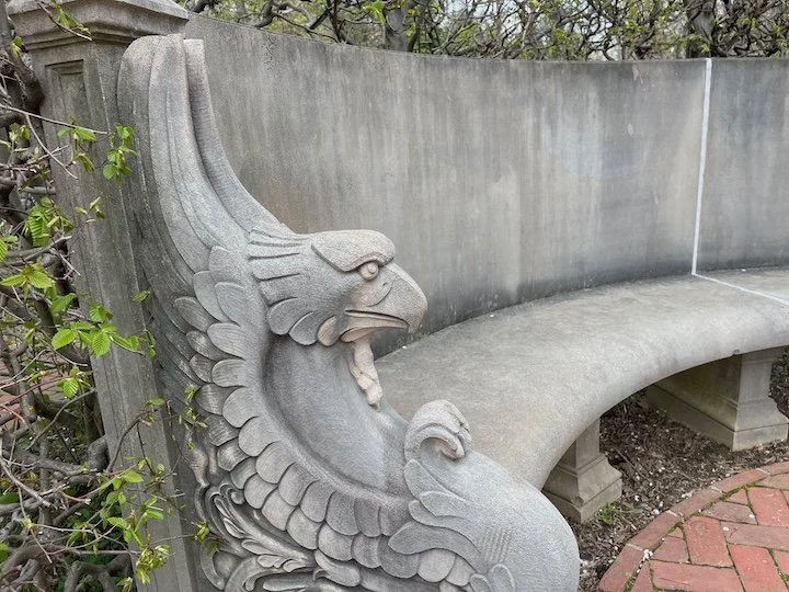 Curved stone garden bench with a carved griffin armrest at Longwood Gardens in Pennsylvania, surrounded by early spring foliage.