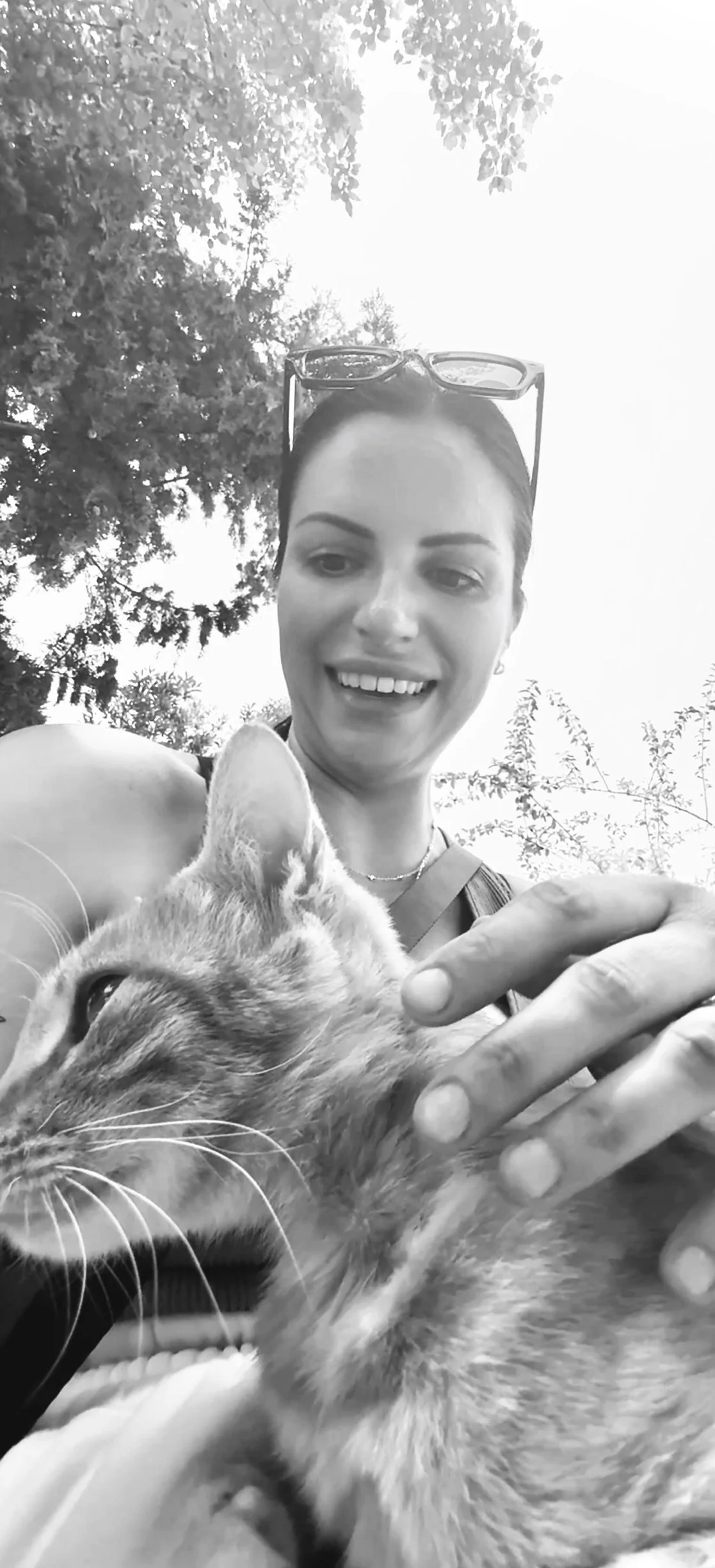 A smiling woman in sunglasses holding a cat outdoors, with trees and sky in the background, in black and white.