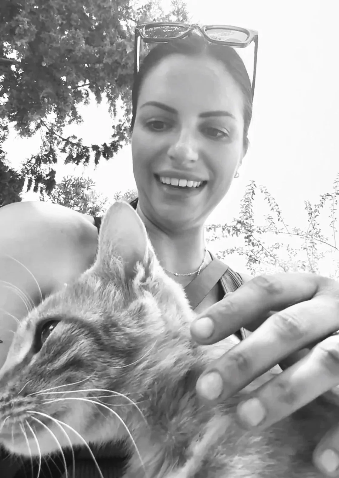 A smiling woman in sunglasses holding a cat outdoors, with trees and sky in the background, in black and white.