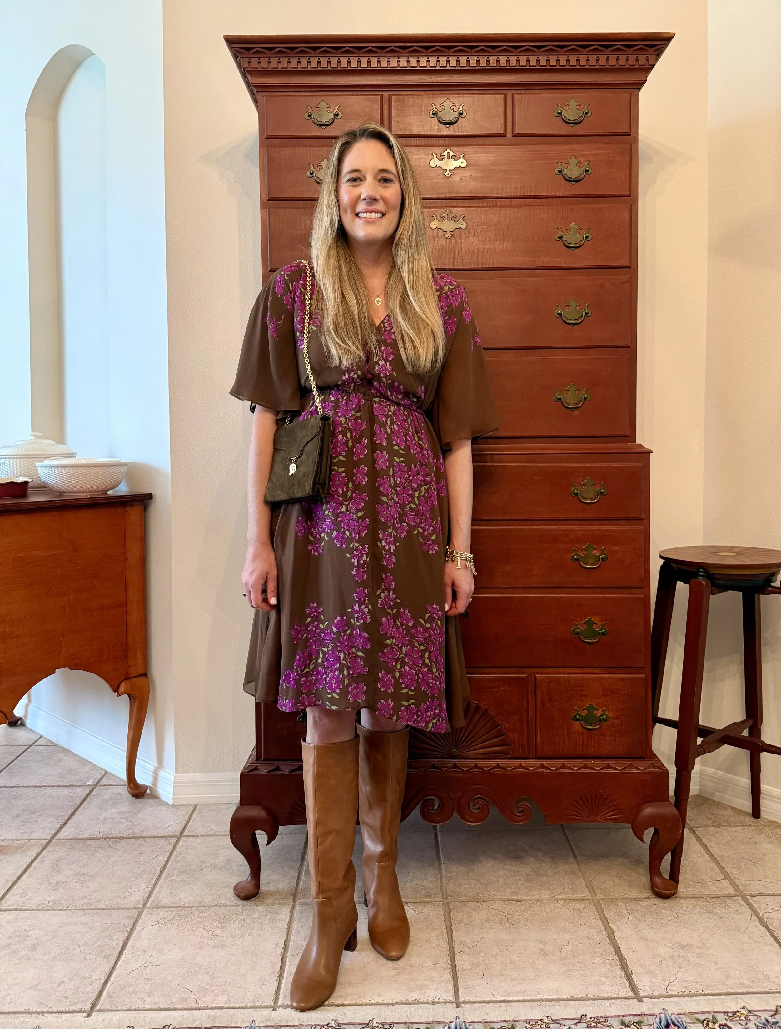 Elizabeth wearing a brown dress with bright pink floral print, belted at the waist, styled with tall camel boots and a small crossbody bag, standing indoors.