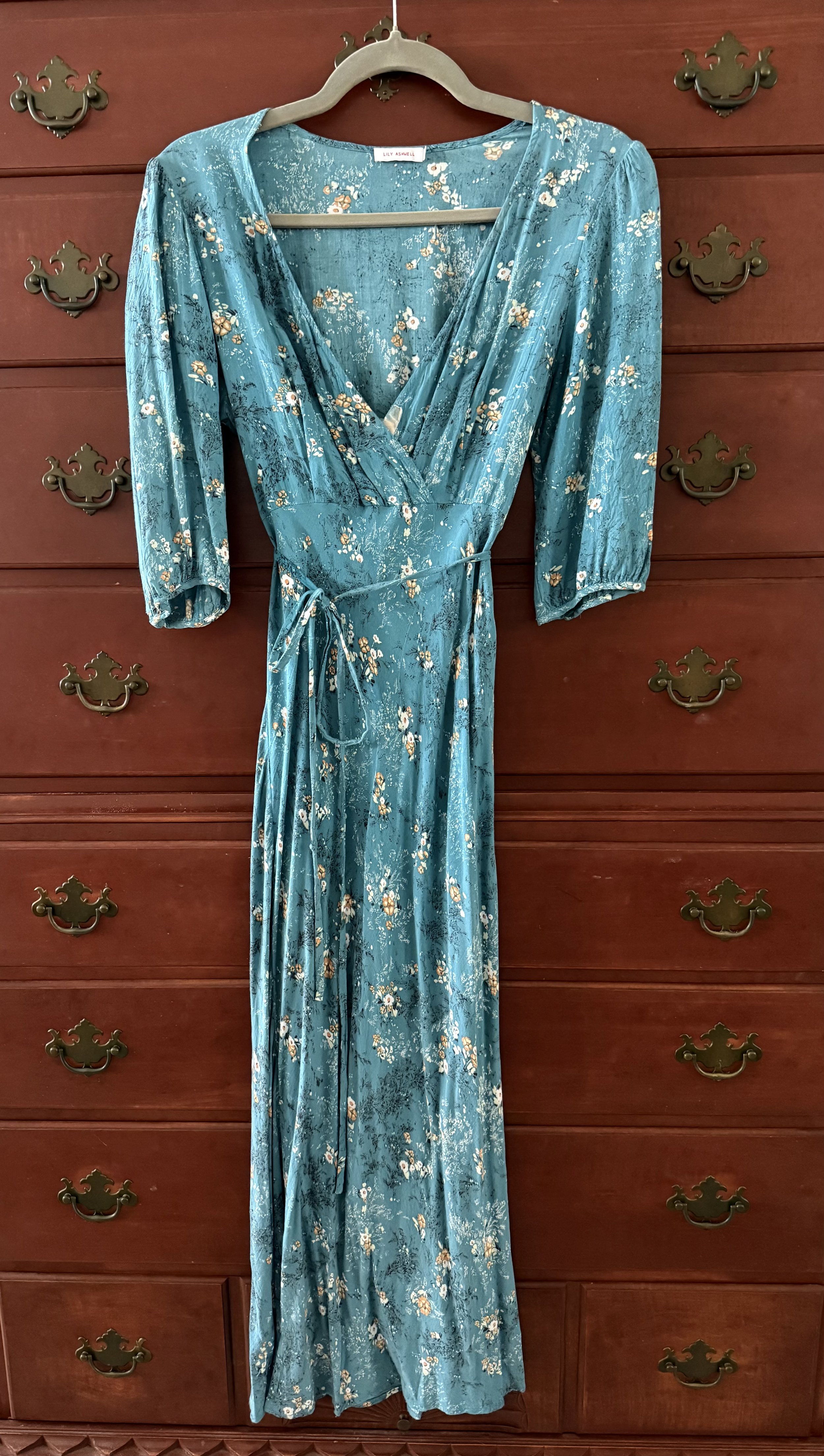 Blue floral wrap dress with short sleeves and tie waist, hanging on a wooden dresser.