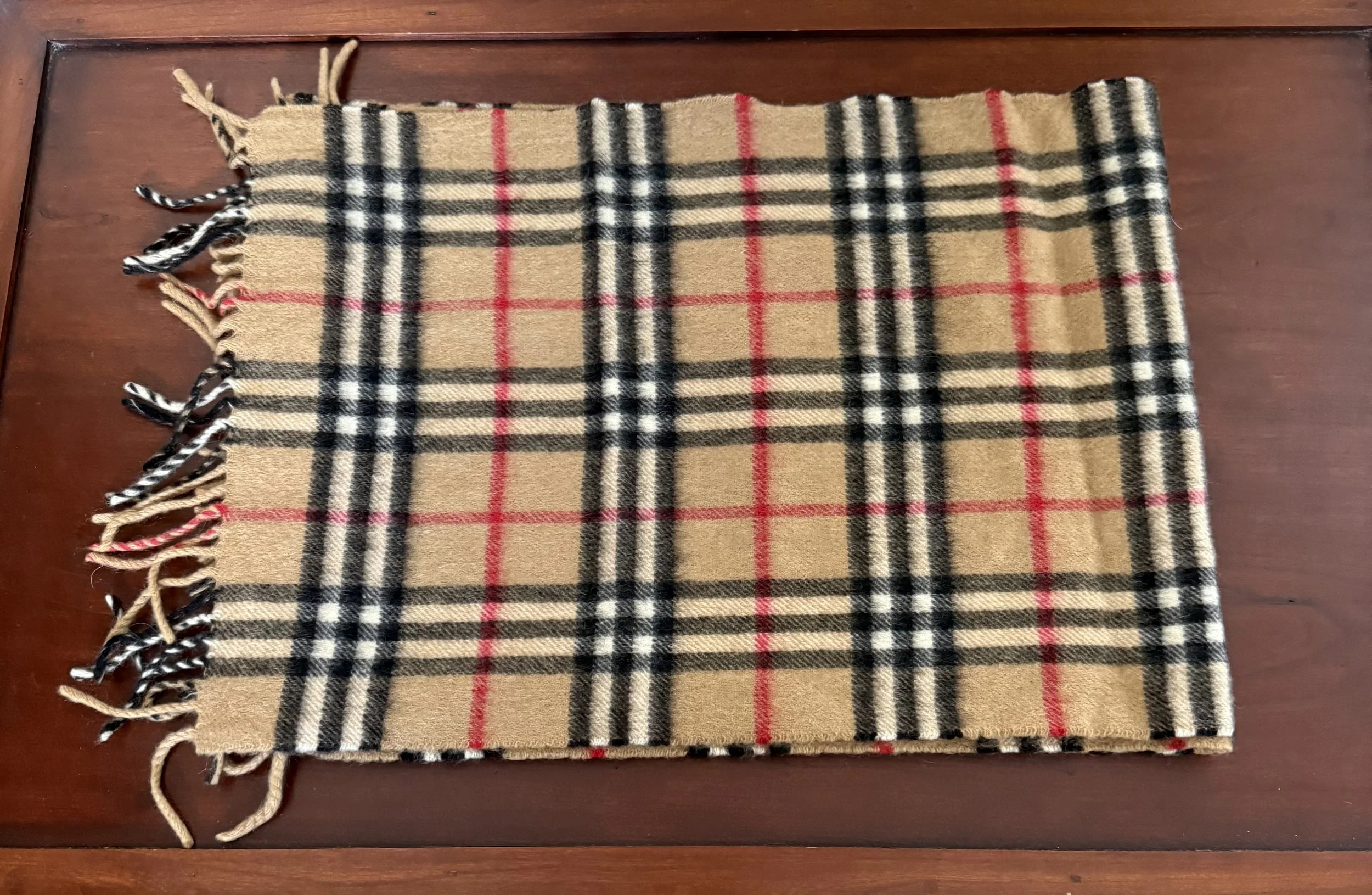 Classic Burberry tan plaid scarf with red, black, and white check pattern, neatly folded on a wooden surface