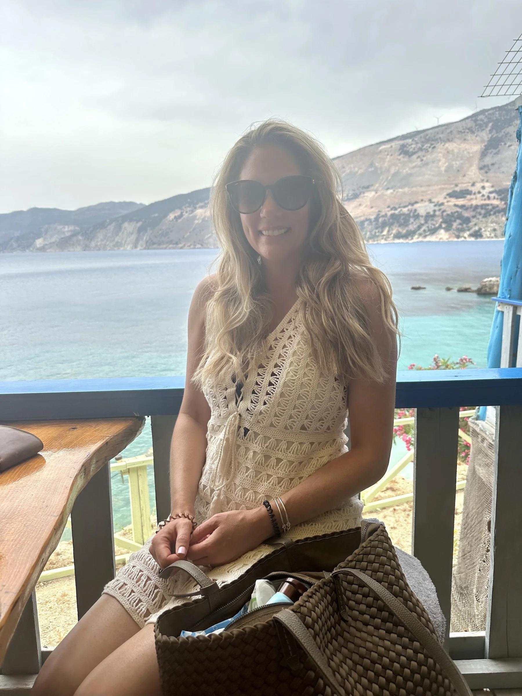 Woman sitting outdoors wearing a cream crochet dress layered over a bikini, styled with minimal accessories in a relaxed tropical setting.