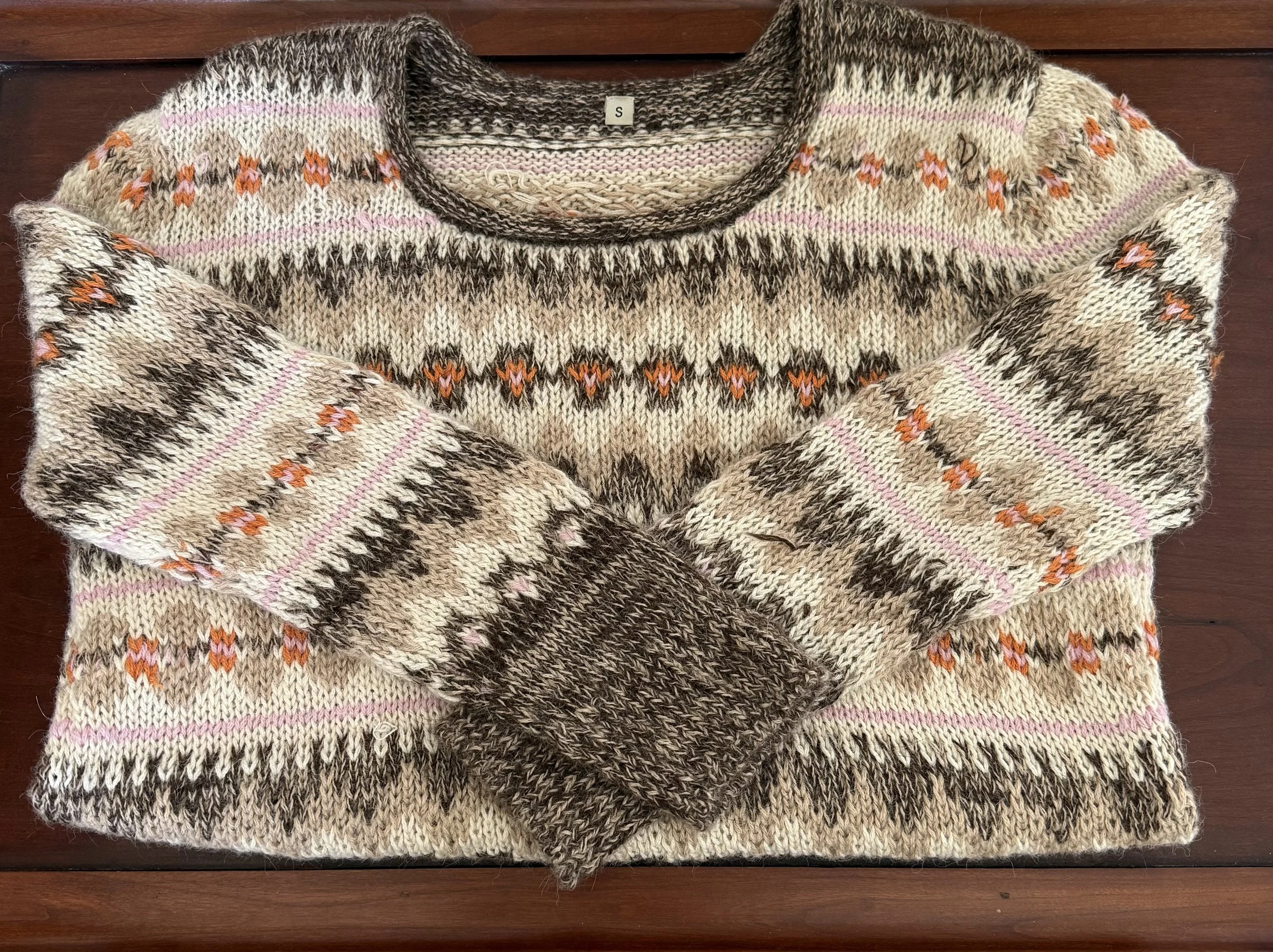 Neutral-toned Fair Isle sweater with pink and orange pattern details, folded neatly on a wooden surface