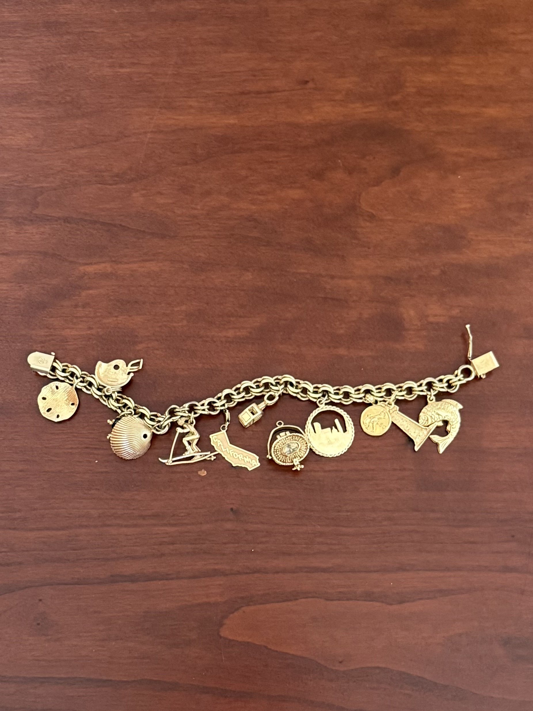 vintage gold charm bracelet with travel-themed charms, including a sand dollar, seashell, sailboat, California state, globe, and other keepsake charms, displayed on a wooden surface
