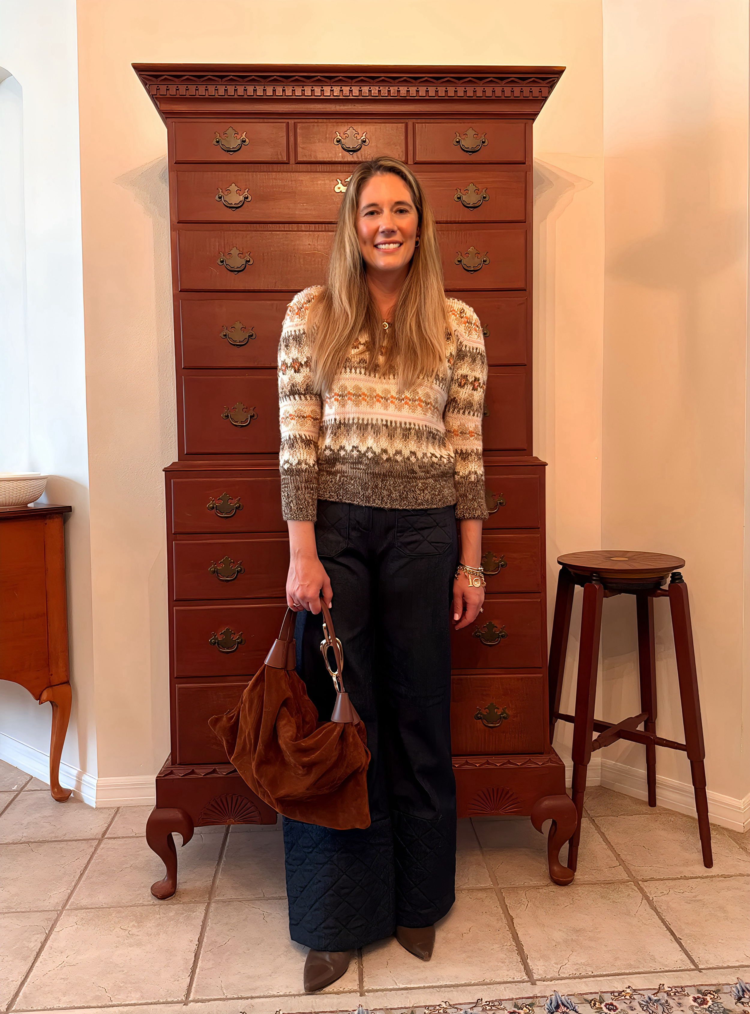 Woman wearing a Lily Ashwell hand-knit Fair Isle sweater with dark wide-leg quilted denim pants, brown suede slouch bag, pointed-toe boots, and a gold charm bracelet, standing indoors against a wooden chest.