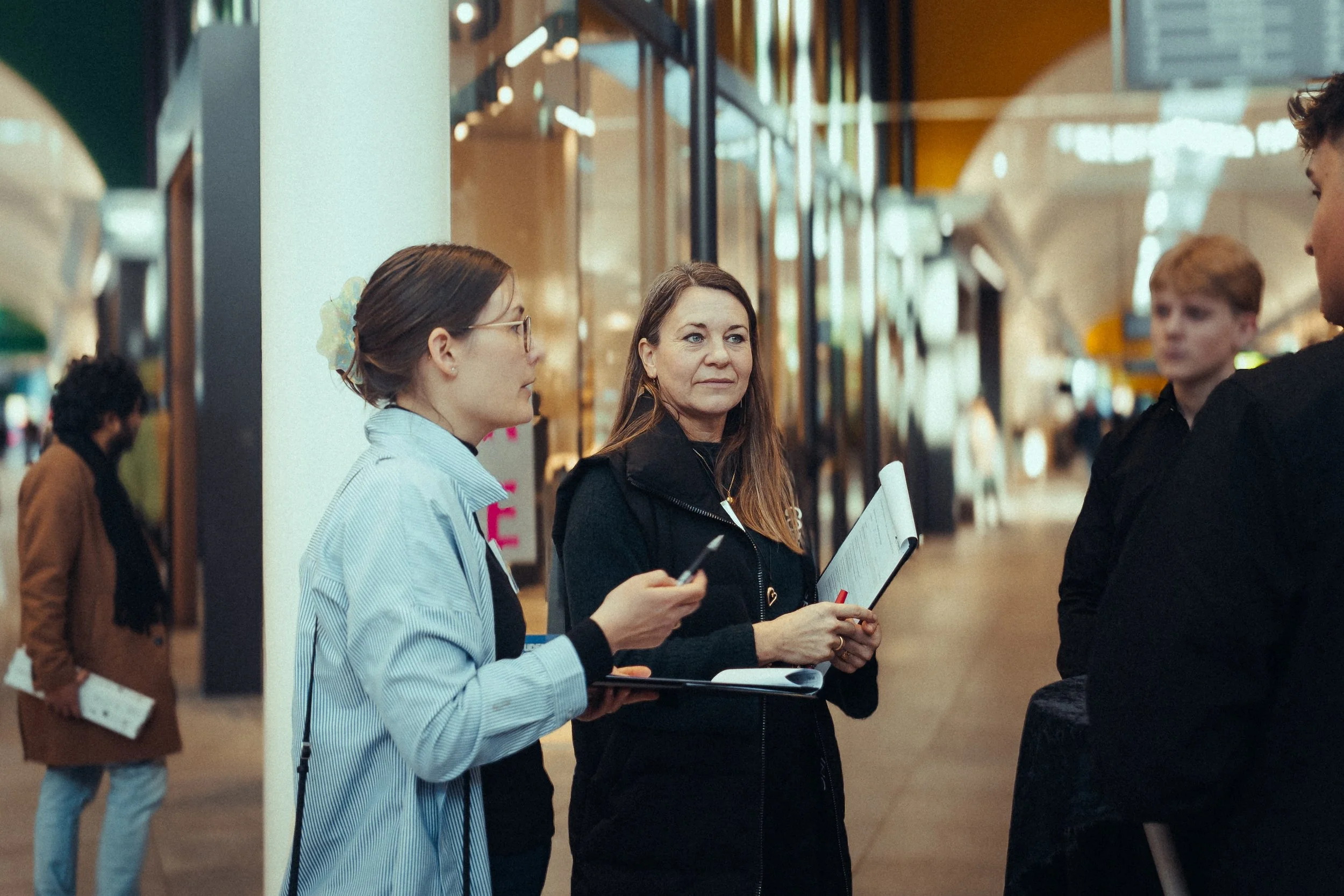 To kvinder står i samtale i et indkøbscenter, en af kvinderne holder en mikrofon og en dokumentmappe, den anden har en telefon.