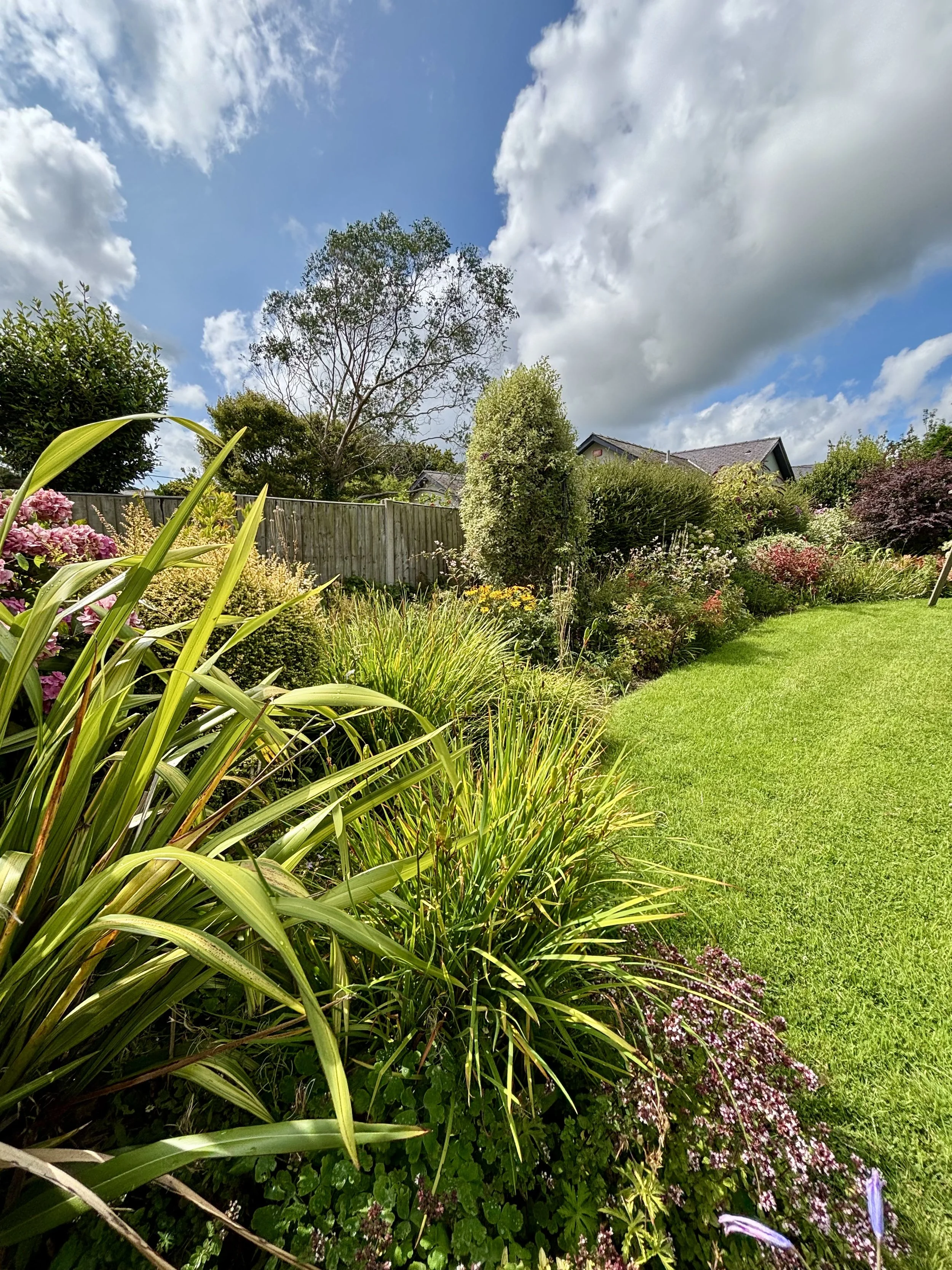 A lush, well-maintained garden with a variety of flowering plants and greenery, a bright green lawn, and mature trees under a partly cloudy blue sky.