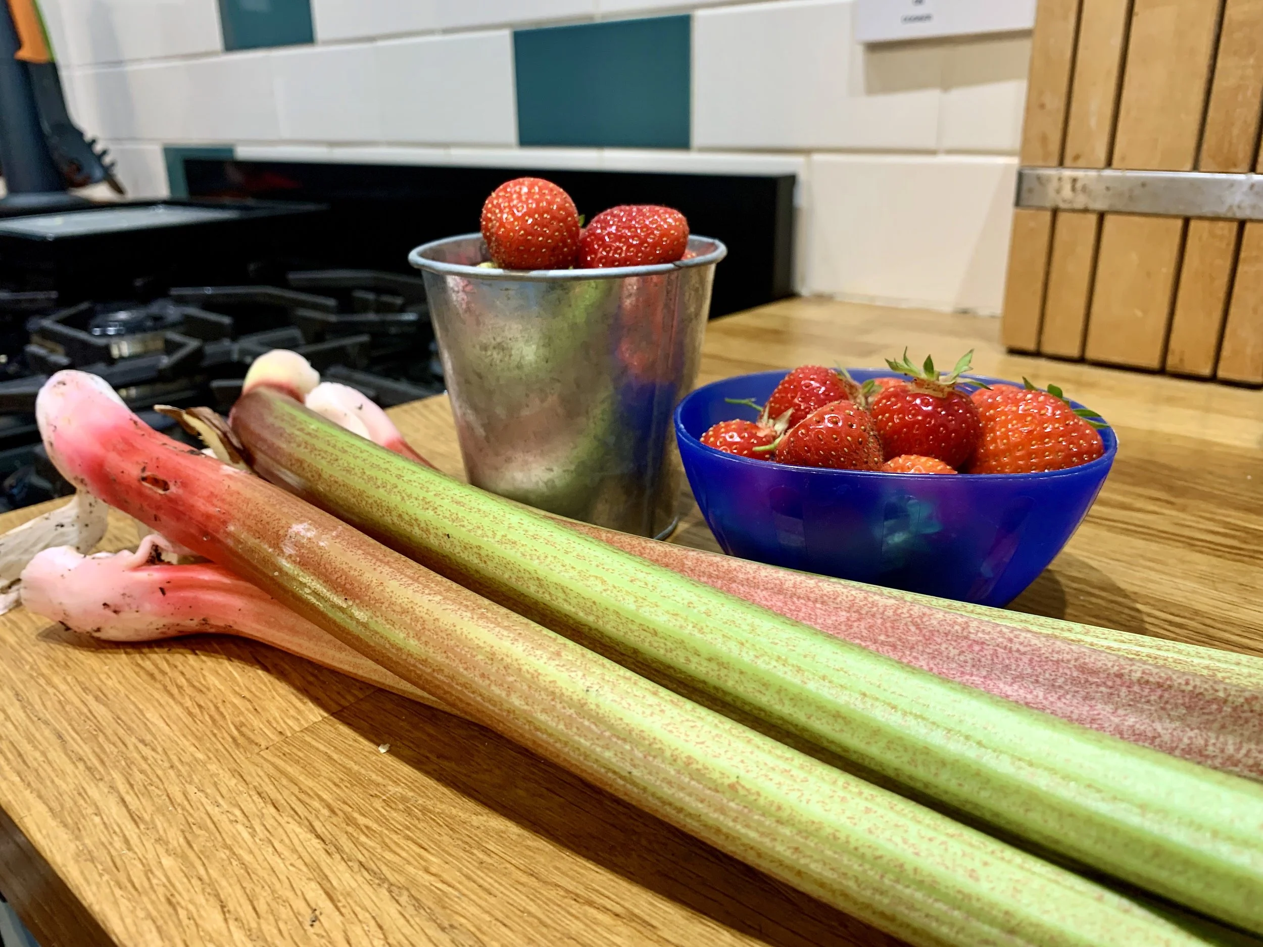 Fresh strawberries in a silver cup, strawberries in a blue glass bowl, and stalks of rhubarb on a wooden countertop in a kitchen.