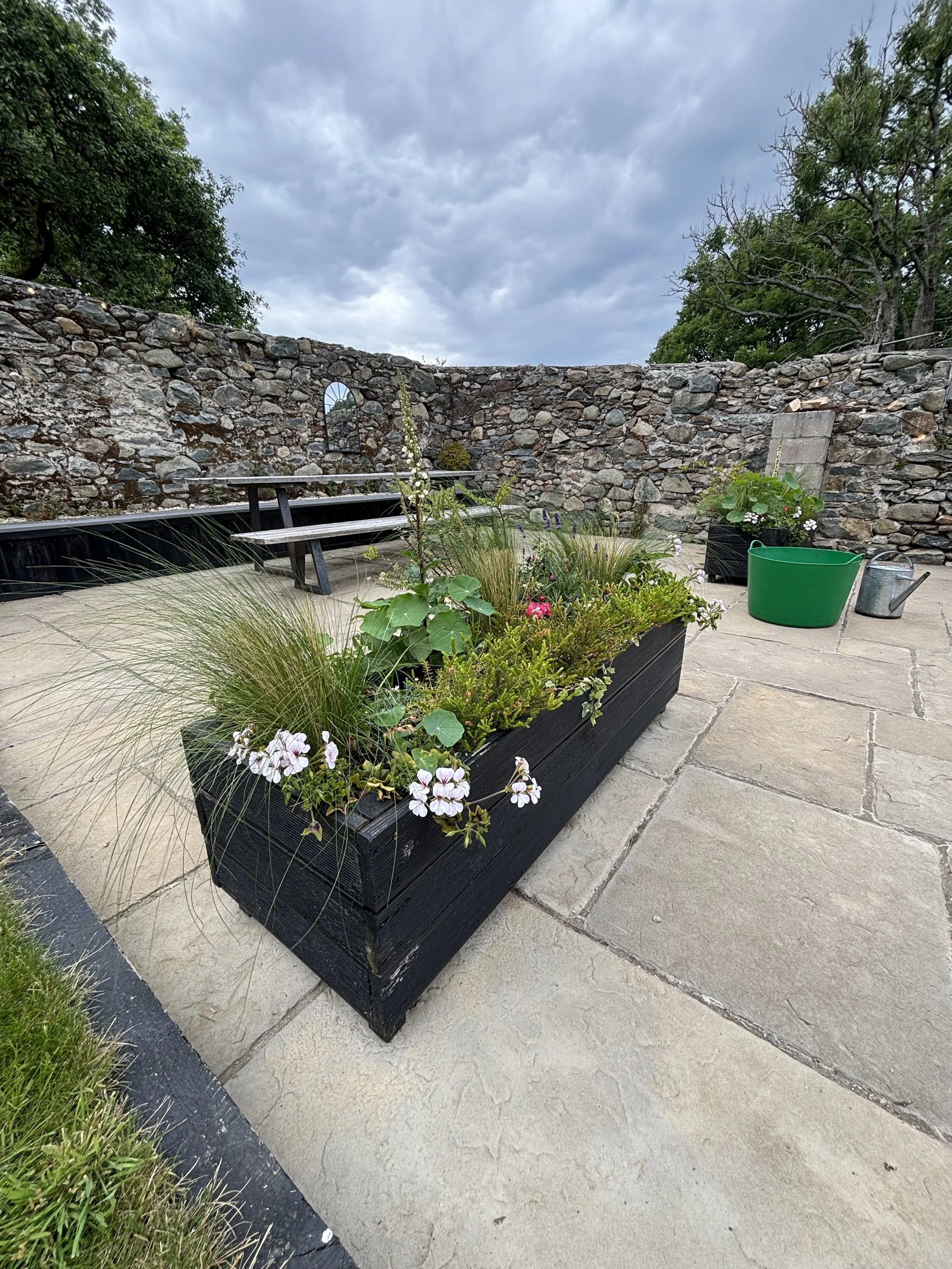 A long black rectangular planter filled with various green plants and flowers, placed on stone patio beside a stone wall and garden tools in a courtyard under a cloudy sky.