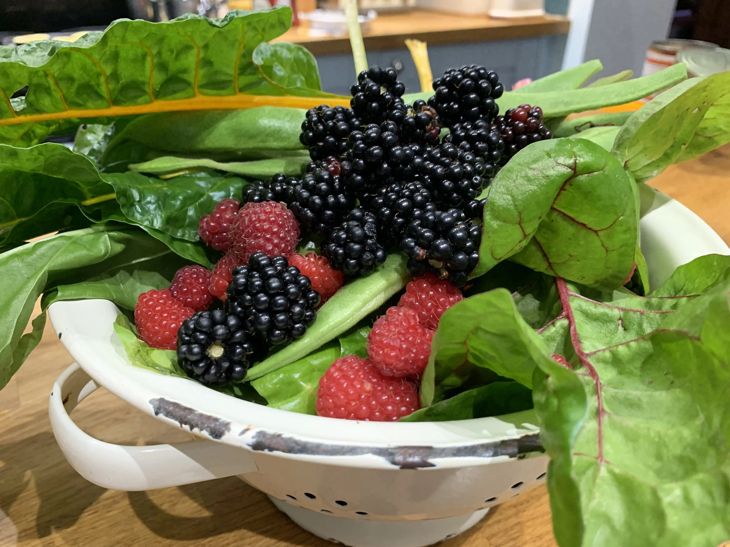 A white ceramic bowl filled with blackberries, raspberries, and various green leafy herbs or vegetables, on a wooden table.