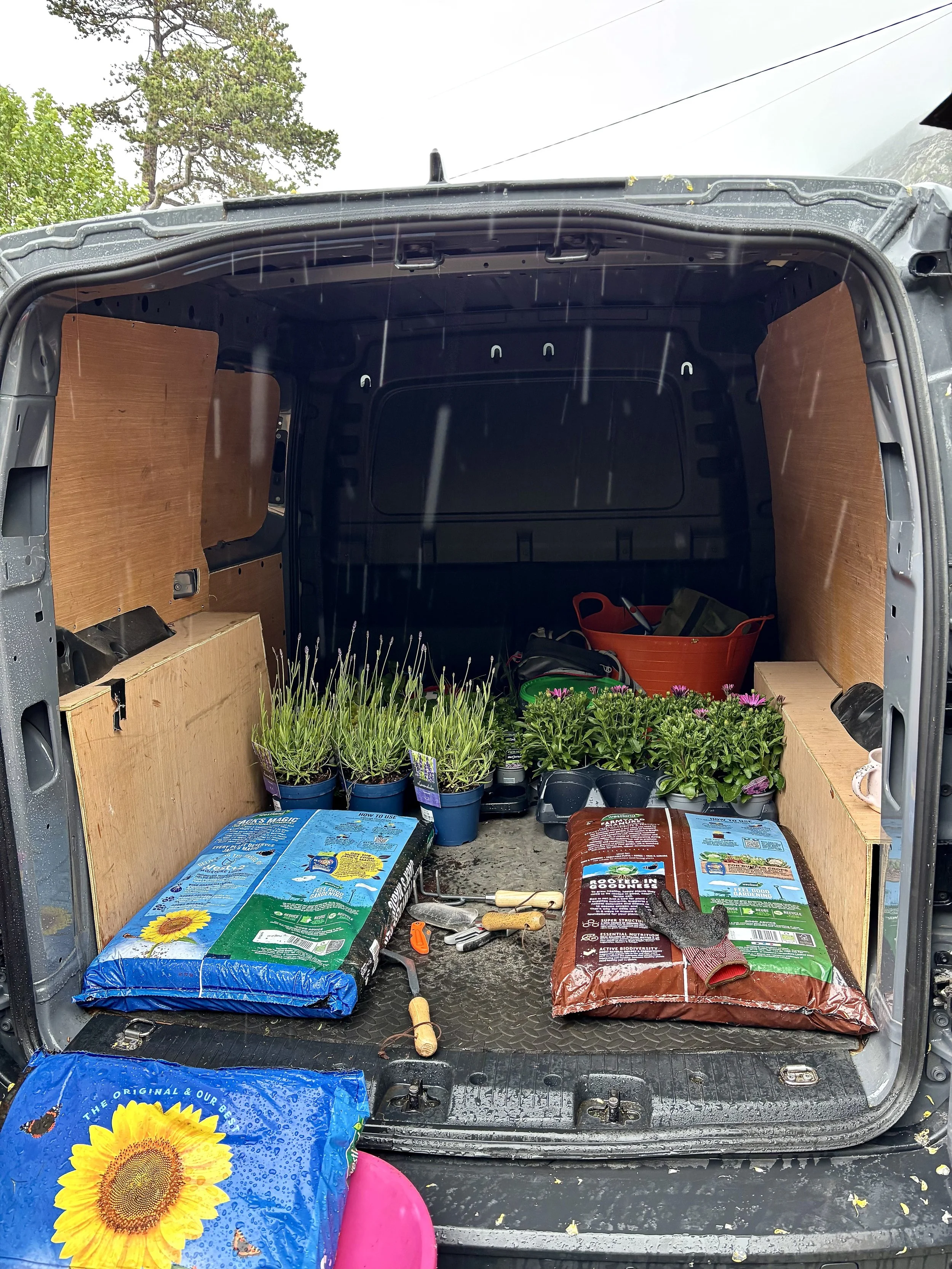 The open back of a vehicle filled with gardening supplies, including bags of soil or mulch, potted plants, gardening tools, and gloves, with a view of trees and cloudy sky outside.