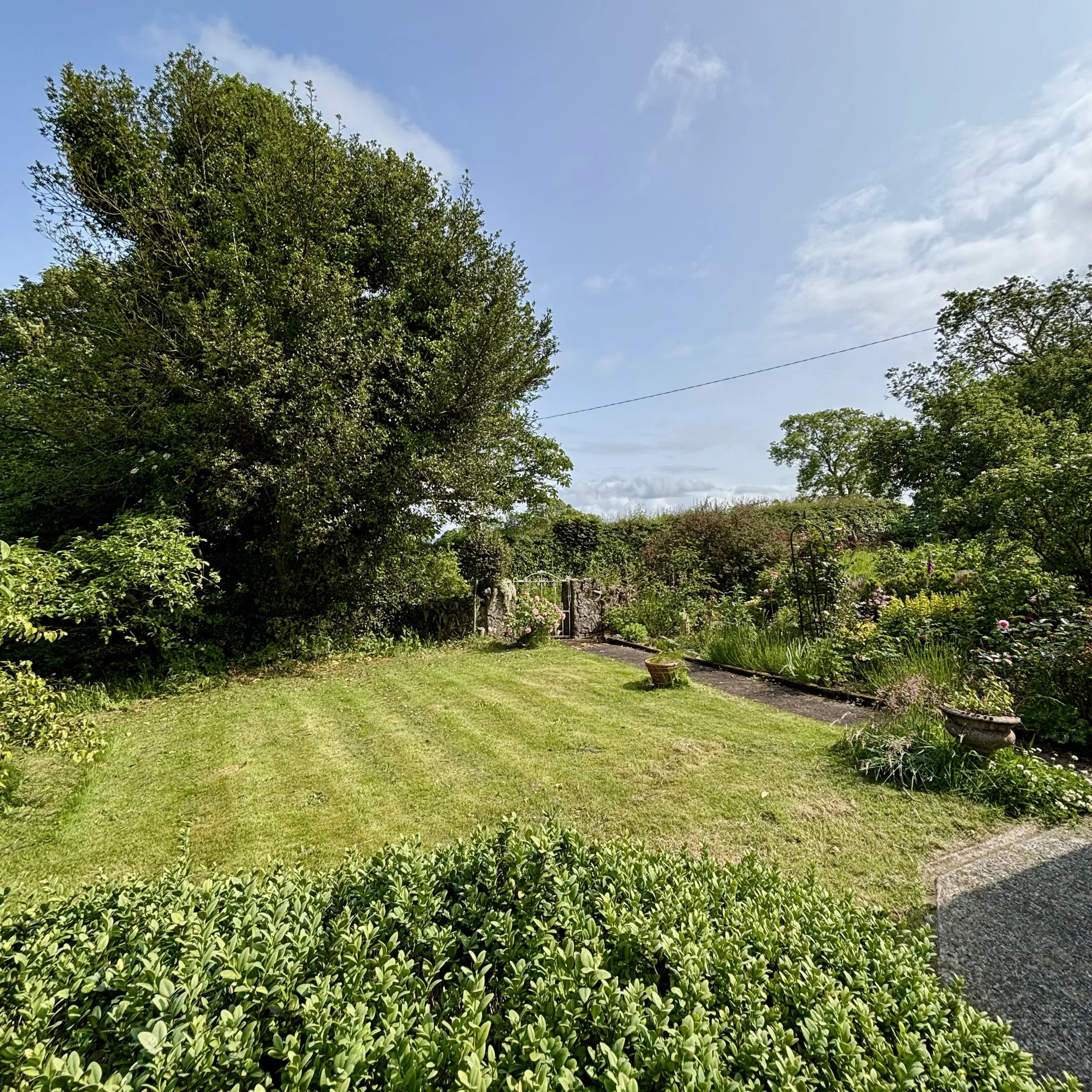 A lush garden with well-maintained green grass, various bushes, and flowering plants, framed by large trees under a partly cloudy sky.