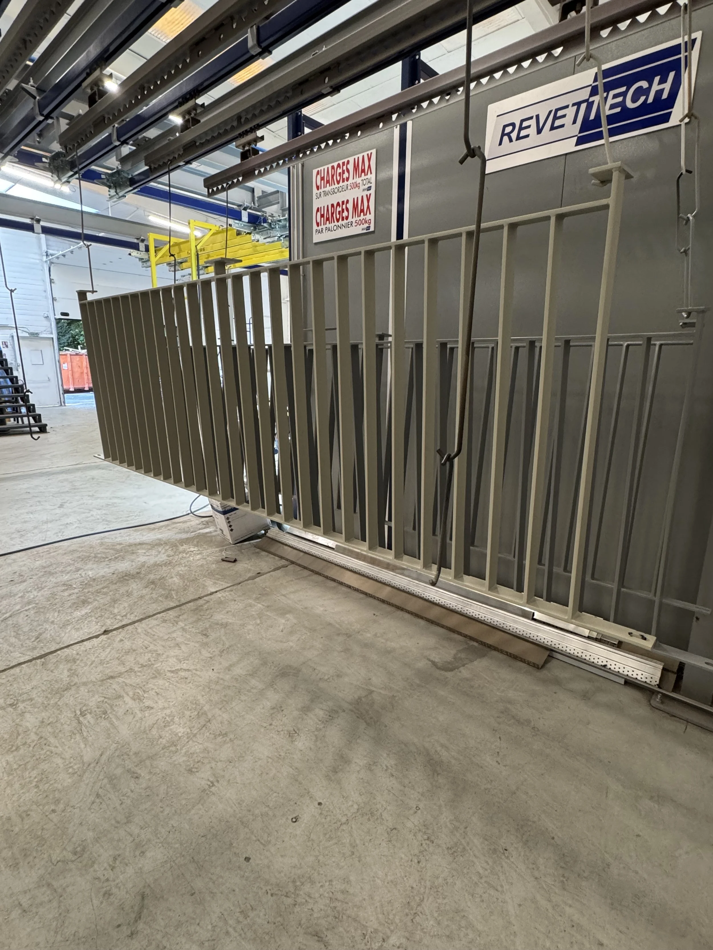 Une porte roulante mécanique avec une barrière de sécurité en métal, située dans un entrepôt ou une usine, avec des panneaux de instructions et de sécurité visibles.