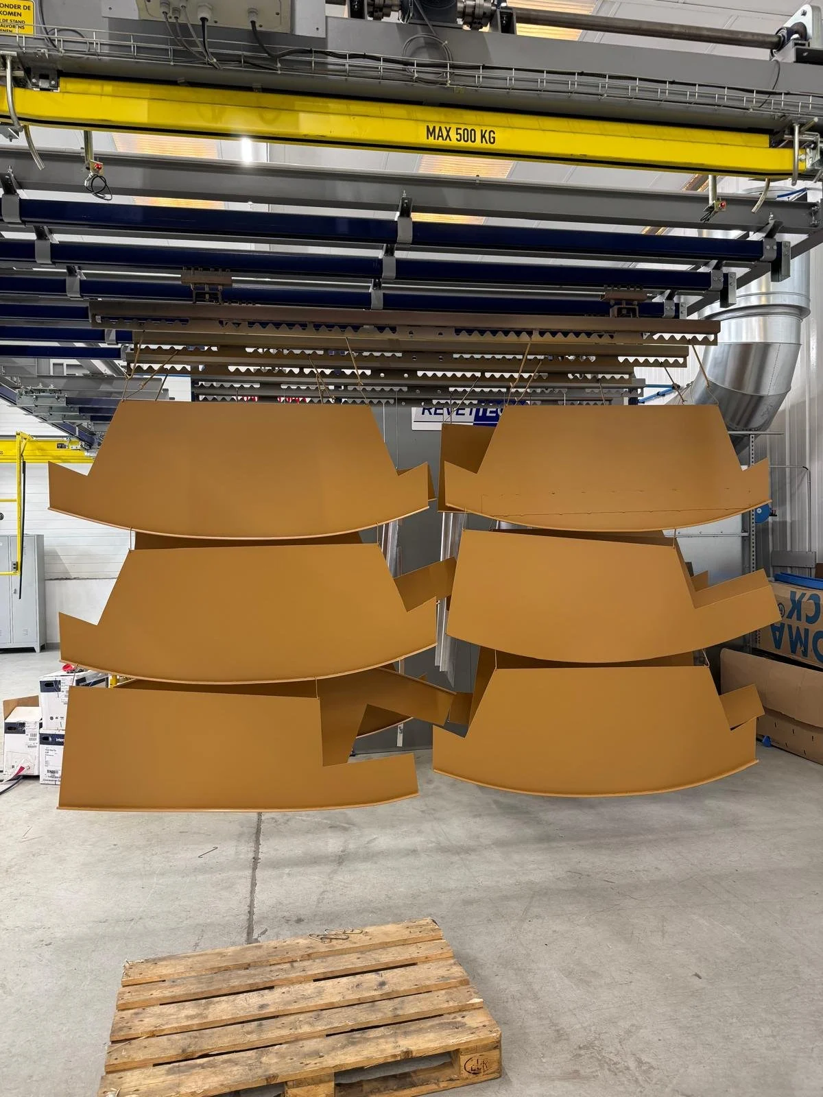 Deux grandes structures métalliques jaunes suspendues dans une usine, avec un dispositif de levage industriel au plafond. Le panneau indique une capacité maximale de 500 kg.