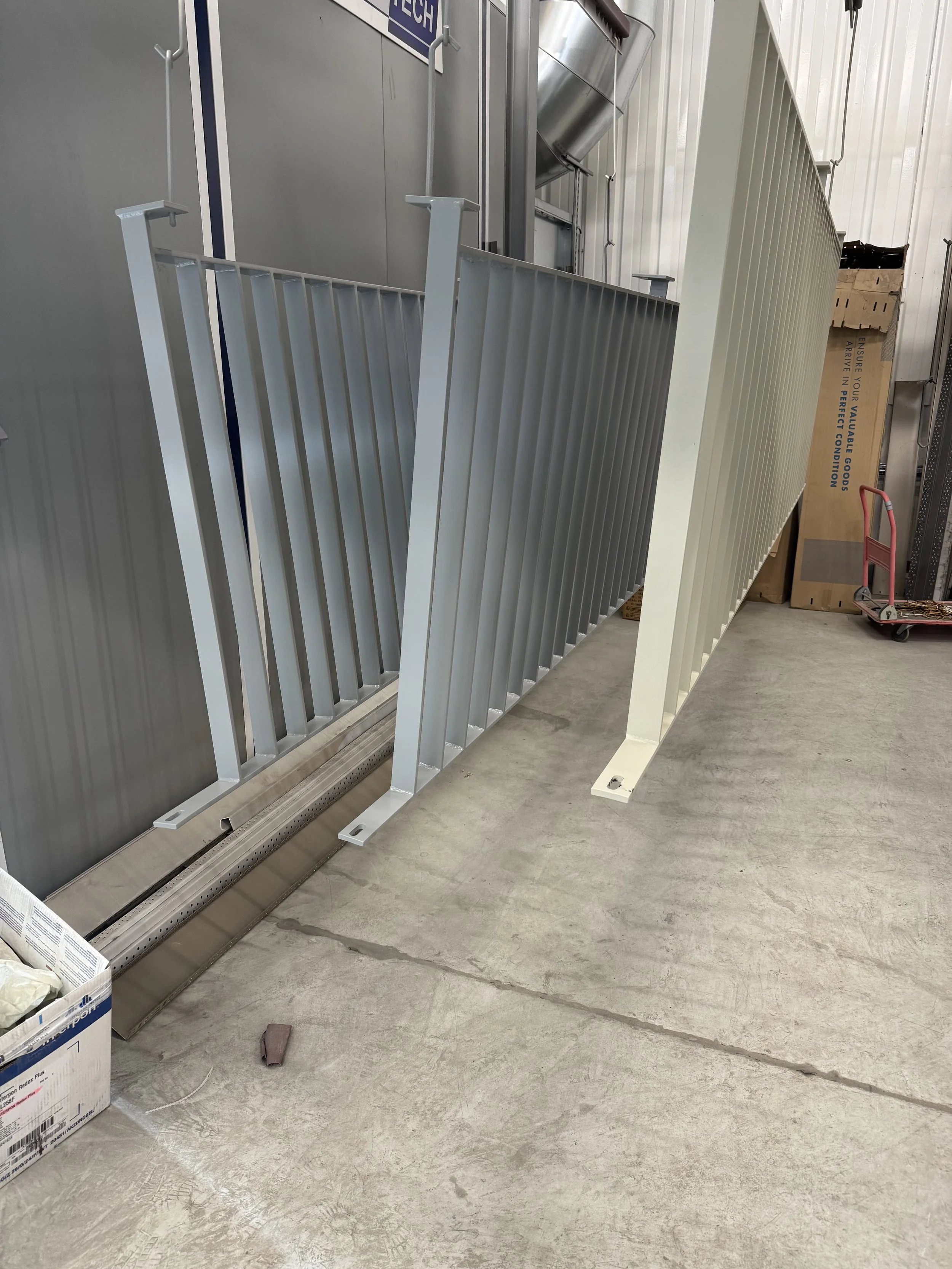 Deux barrières métalliques blanches et une barrière grise dans un entrepôt ou une zone industrielle, avec des cartons et un chariot en arrière-plan.