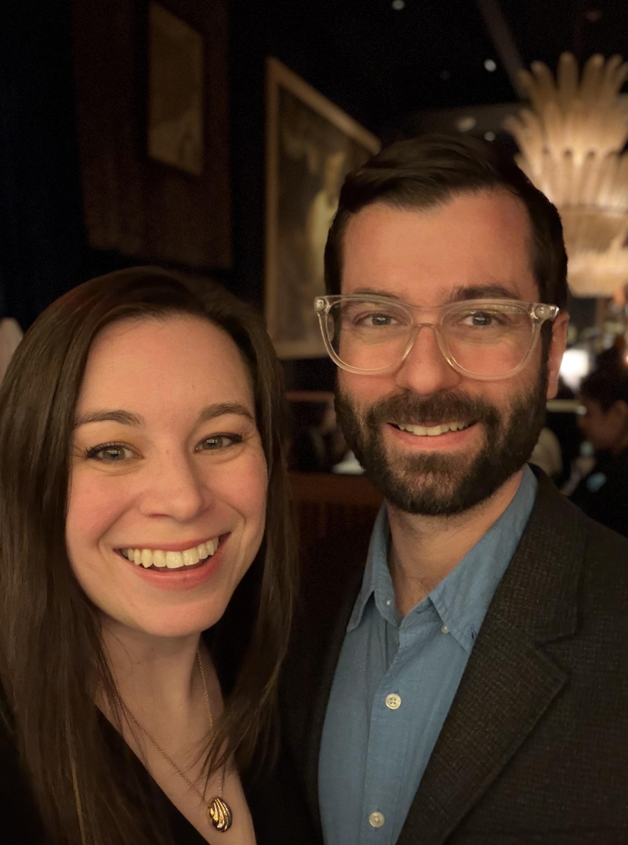 A man and woman smiling and posing for a photo in a dimly lit indoor setting, possibly a restaurant or bar.