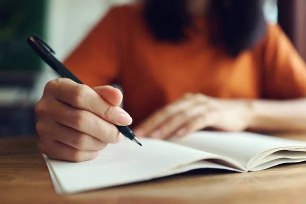 Person writing in a notebook with a black pen, focus on hand and pen, blurred background.