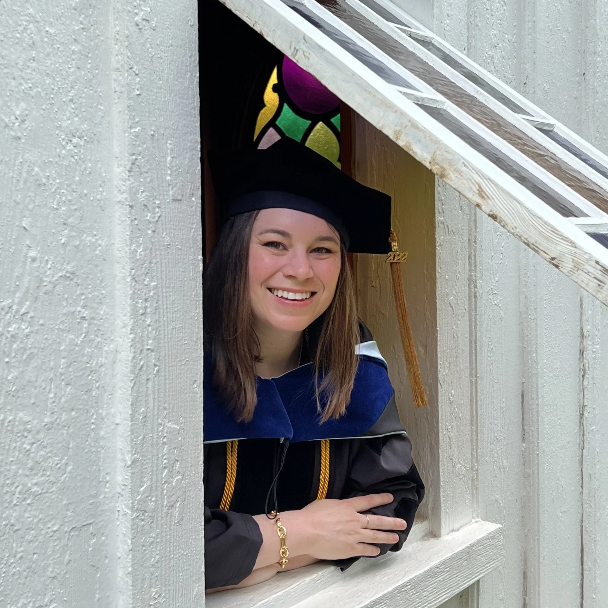 Young woman in graduation gown and cap smiling and leaning on a window sill.