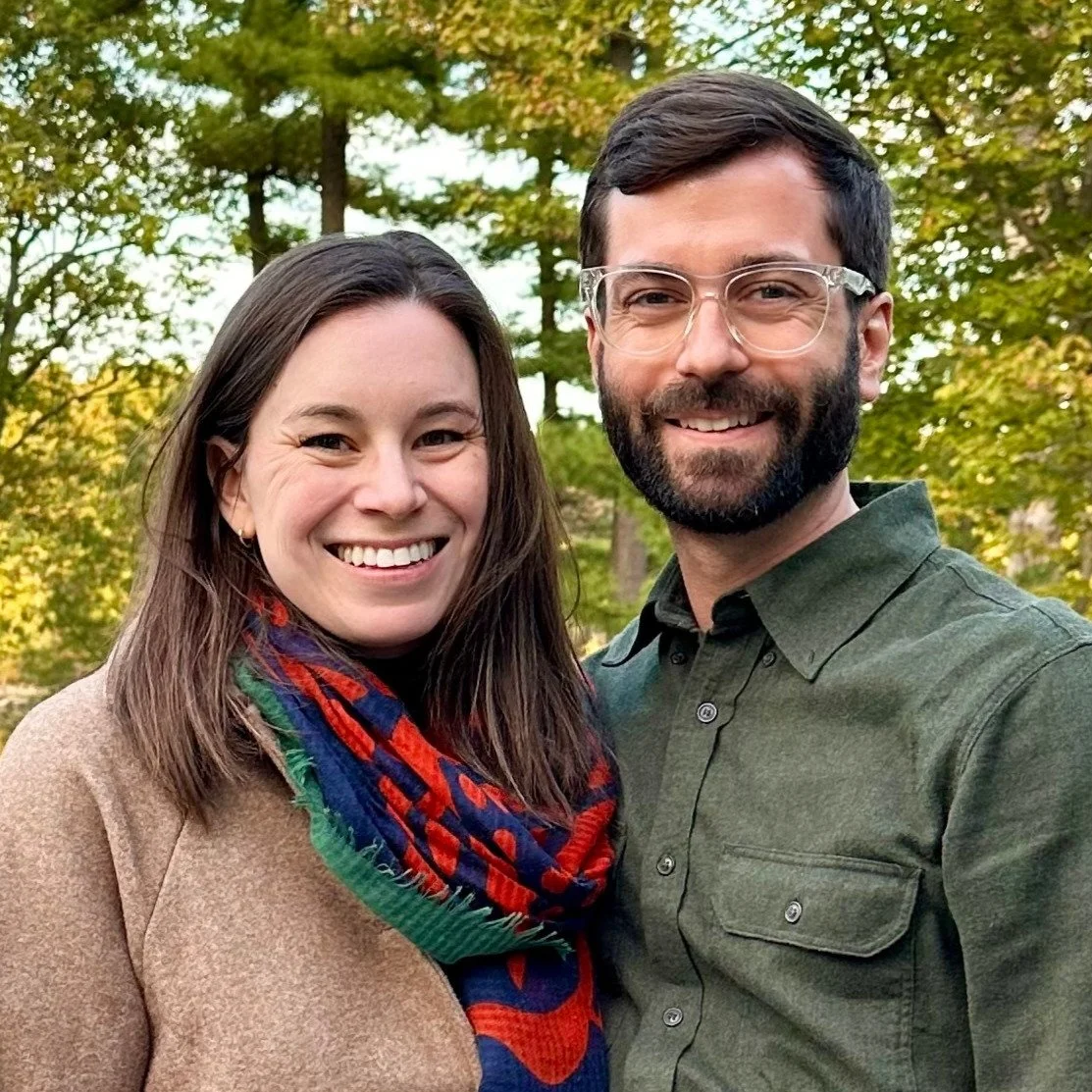 A smiling couple outdoors with trees and fall foliage in the background.