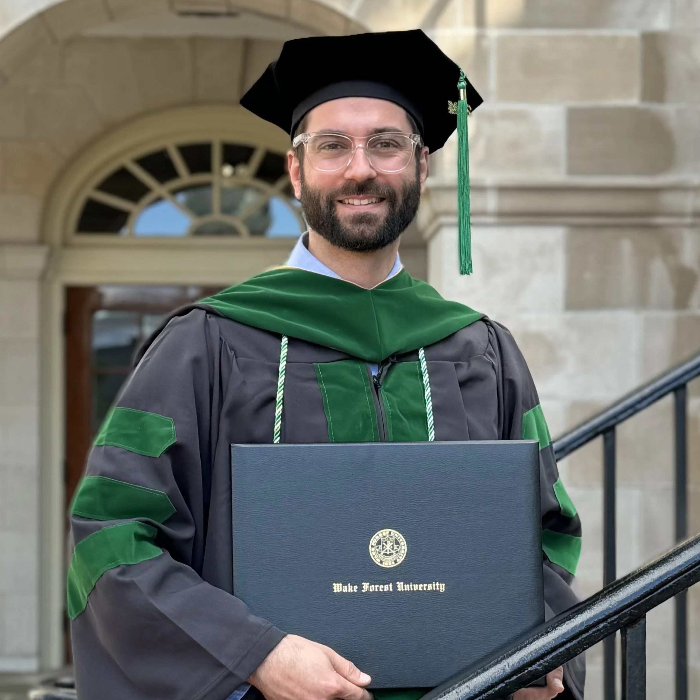 A man in graduation attire with a green and black gown and cap, holding a diploma from Wake Forest University, standing in front of a historic building.