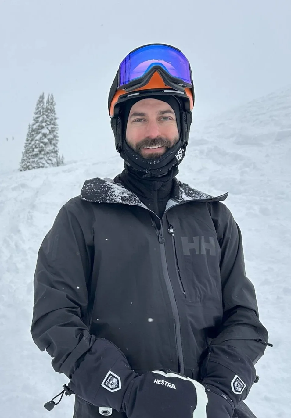 A man in winter gear, including a black jacket, gloves, and ski helmet with goggles, standing outdoors in a snowy landscape with snow-covered trees.