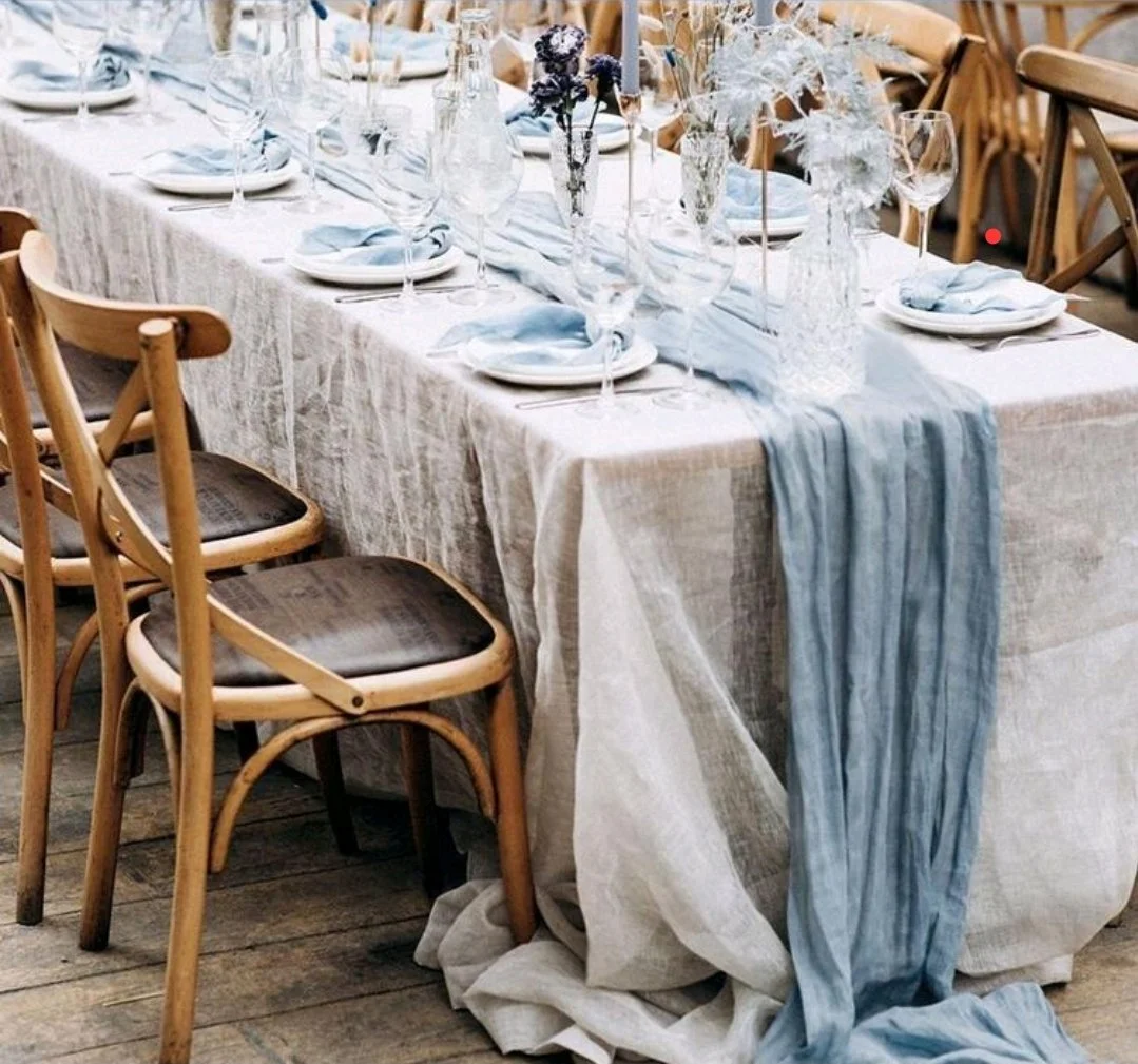 A long dining table decorated for a formal event, with white tablecloth, blue fabric runner, glassware, plates, and floral centerpieces, surrounded by wooden chairs.
