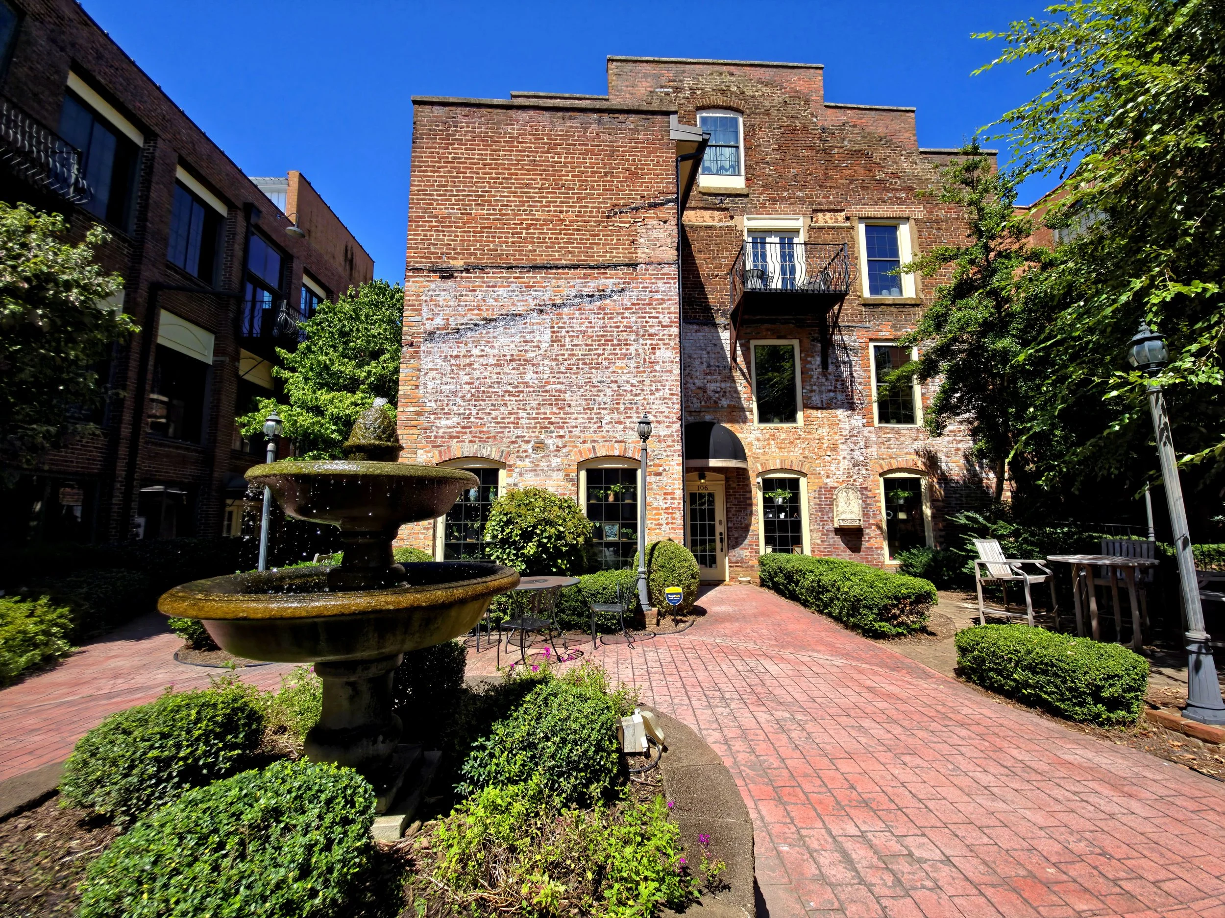 Brick building with multiple windows and a small balcony, surrounded by green trees, shrubs, and a garden with a stone fountain, benches, and a brick pathway under a clear blue sky.