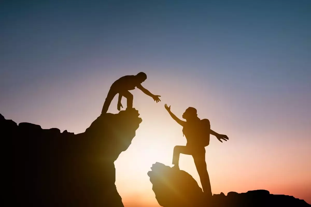 Silhouette of two hikers reaching out to help each other on mountain cliffs during sunset