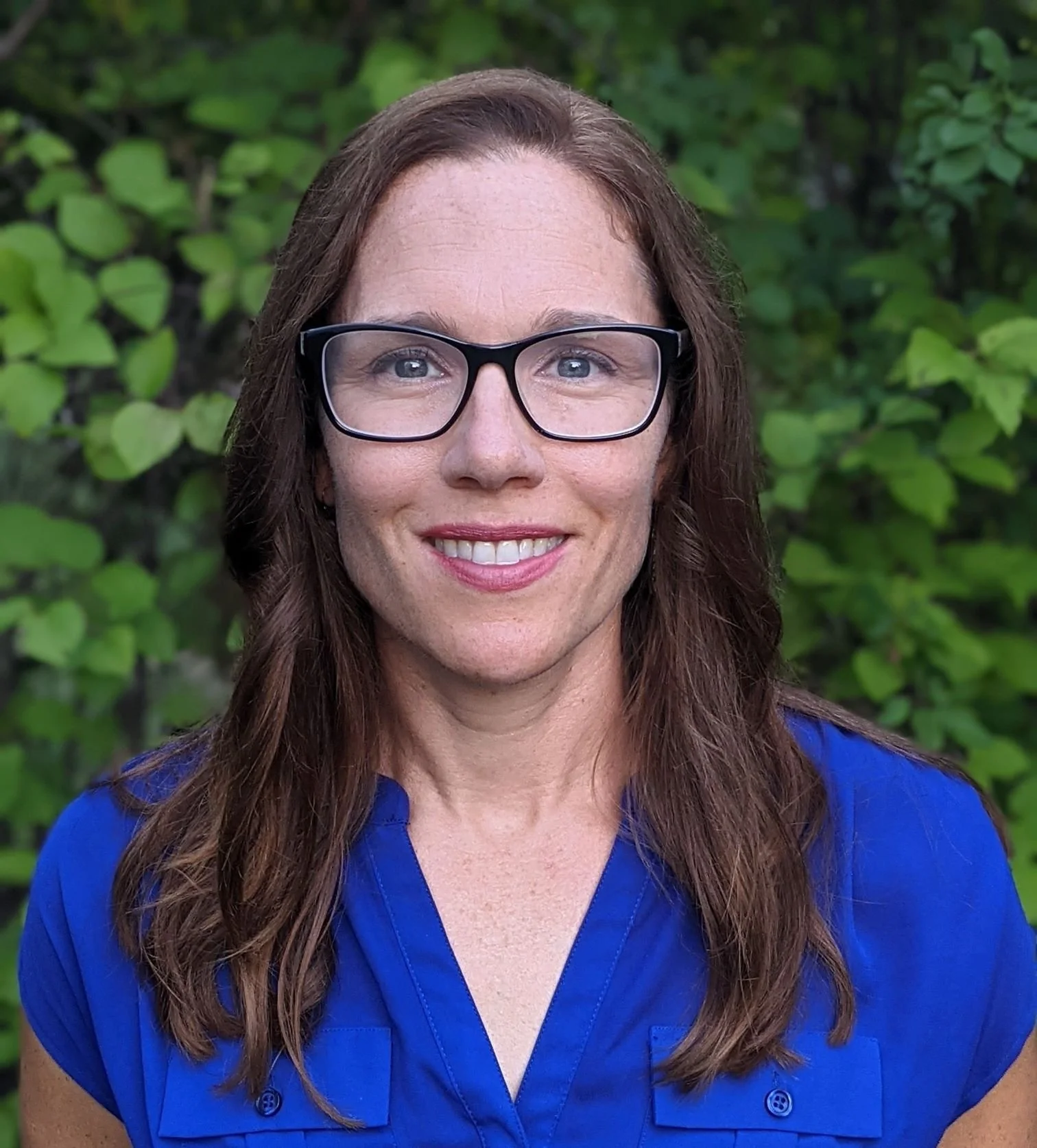 Portrait of a woman with long brown hair, wearing glasses and a blue shirt, standing outdoors in front of green foliage.