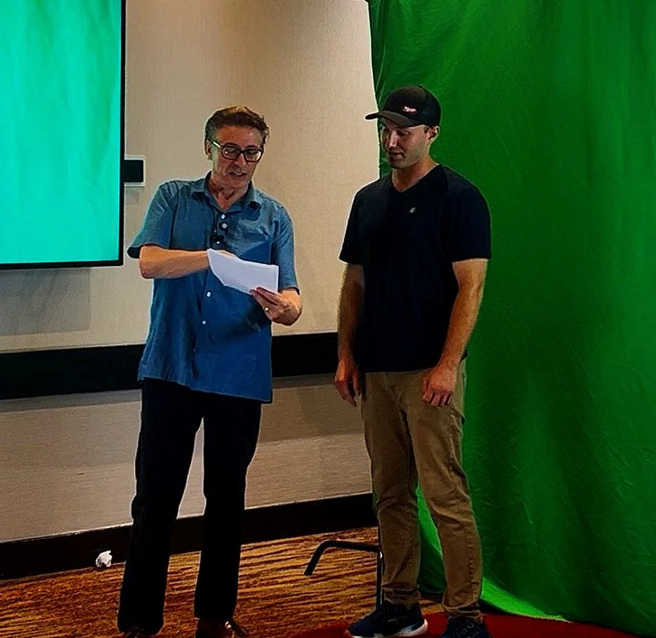 Two men standing indoors, one holding a piece of paper, with a green screen behind them and a large screen on the wall.