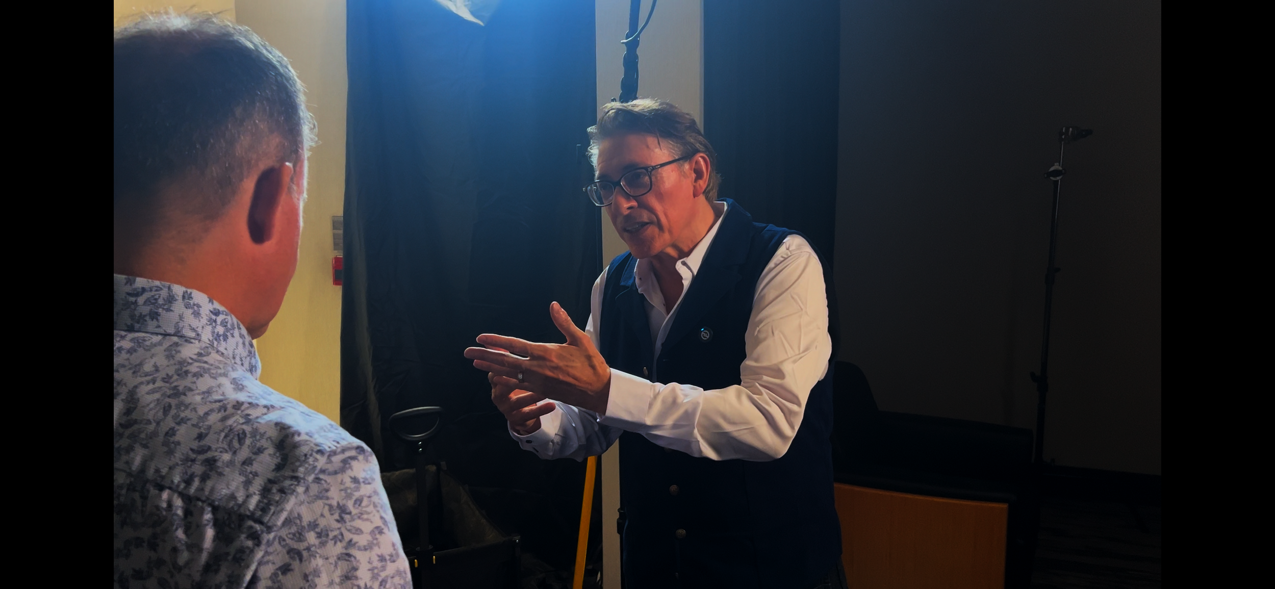 Two men engaged in a conversation, one is gesturing with his hands while speaking, and the other is listening, in an indoor setting with a dark background, a black curtain, and studio lighting equipment.