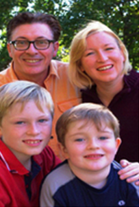 A smiling family of four outdoors, with two adults and two children, surrounded by green trees.