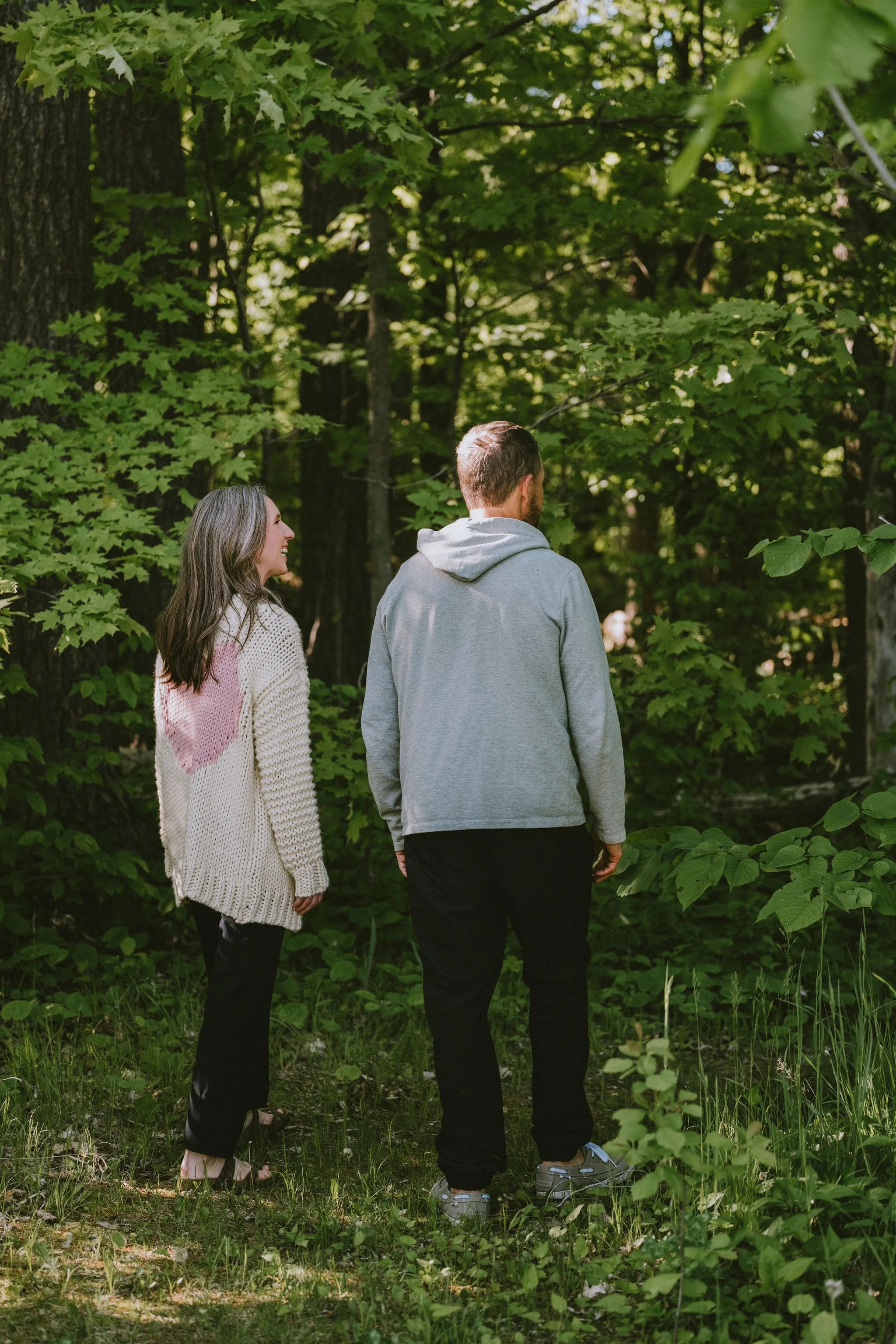 A man and woman walking through a forest, surrounded by green leaves and trees, with the woman smiling and looking at the man.