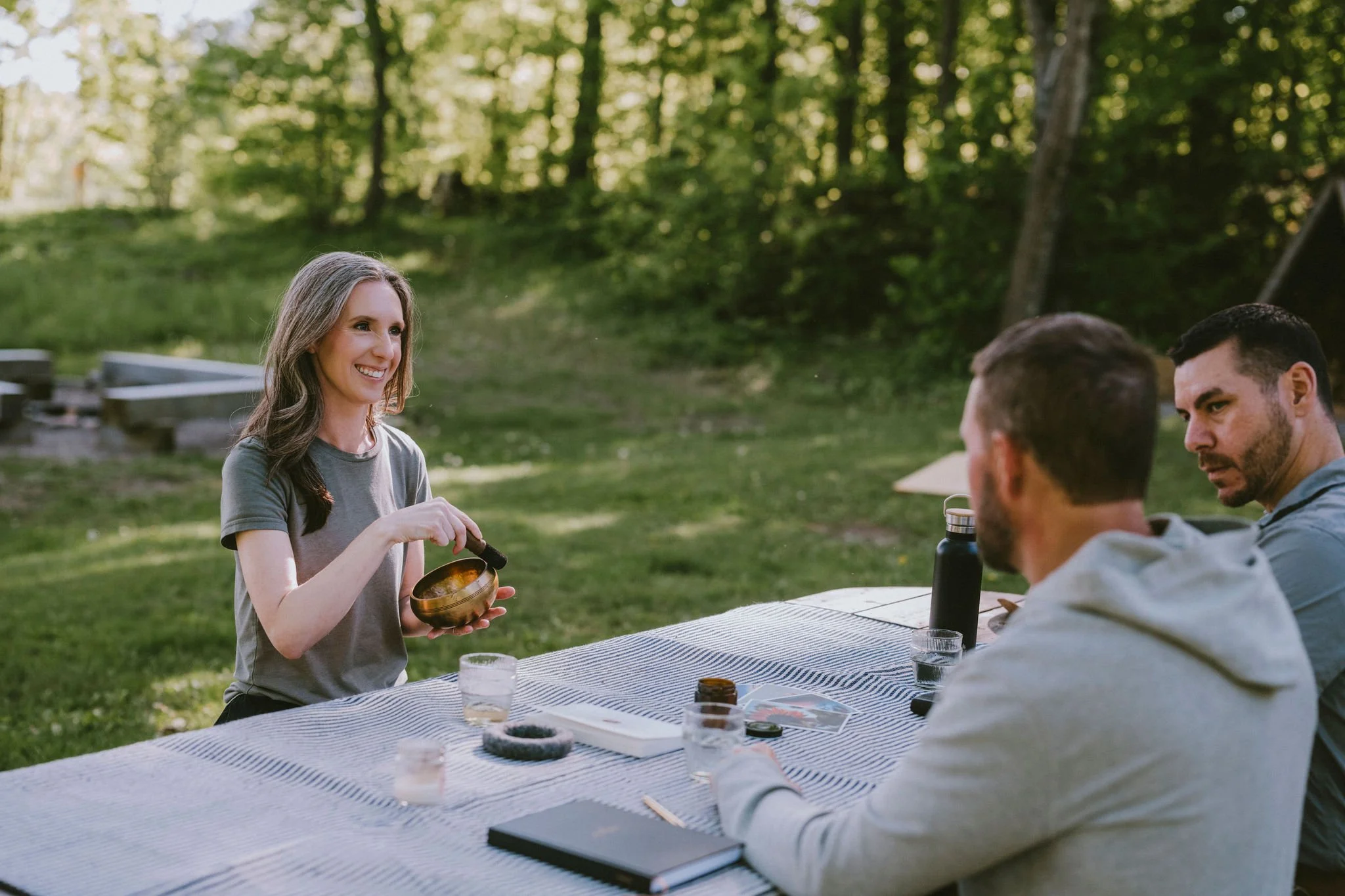 Three people sit at a table outdoors, with a woman showing a singing bowl to two men during daytime.