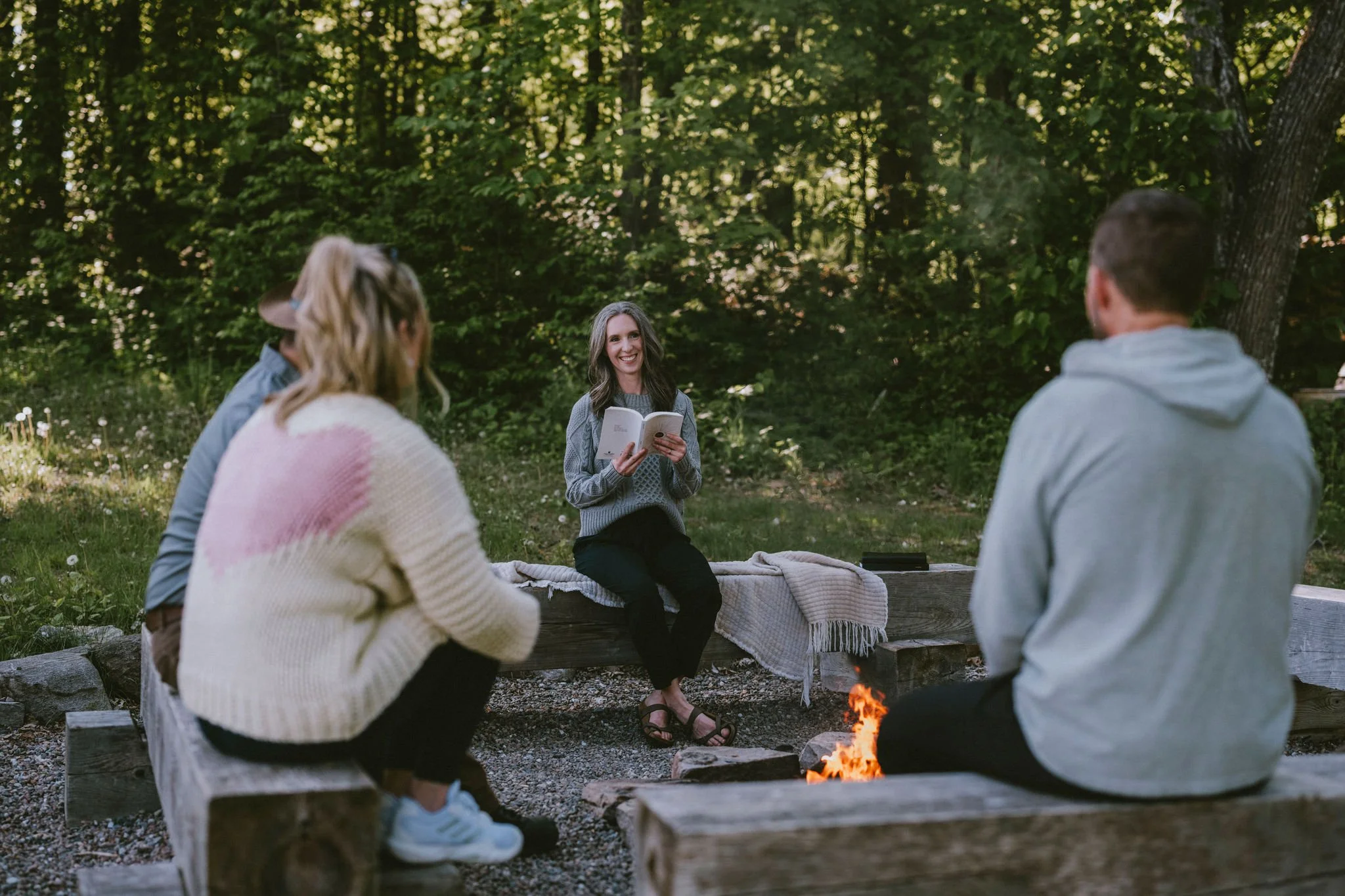 A group of people sitting around a campfire in a wooded outdoor setting. One woman is sitting on a wooden log, reading a book and smiling, while others sit nearby listening.