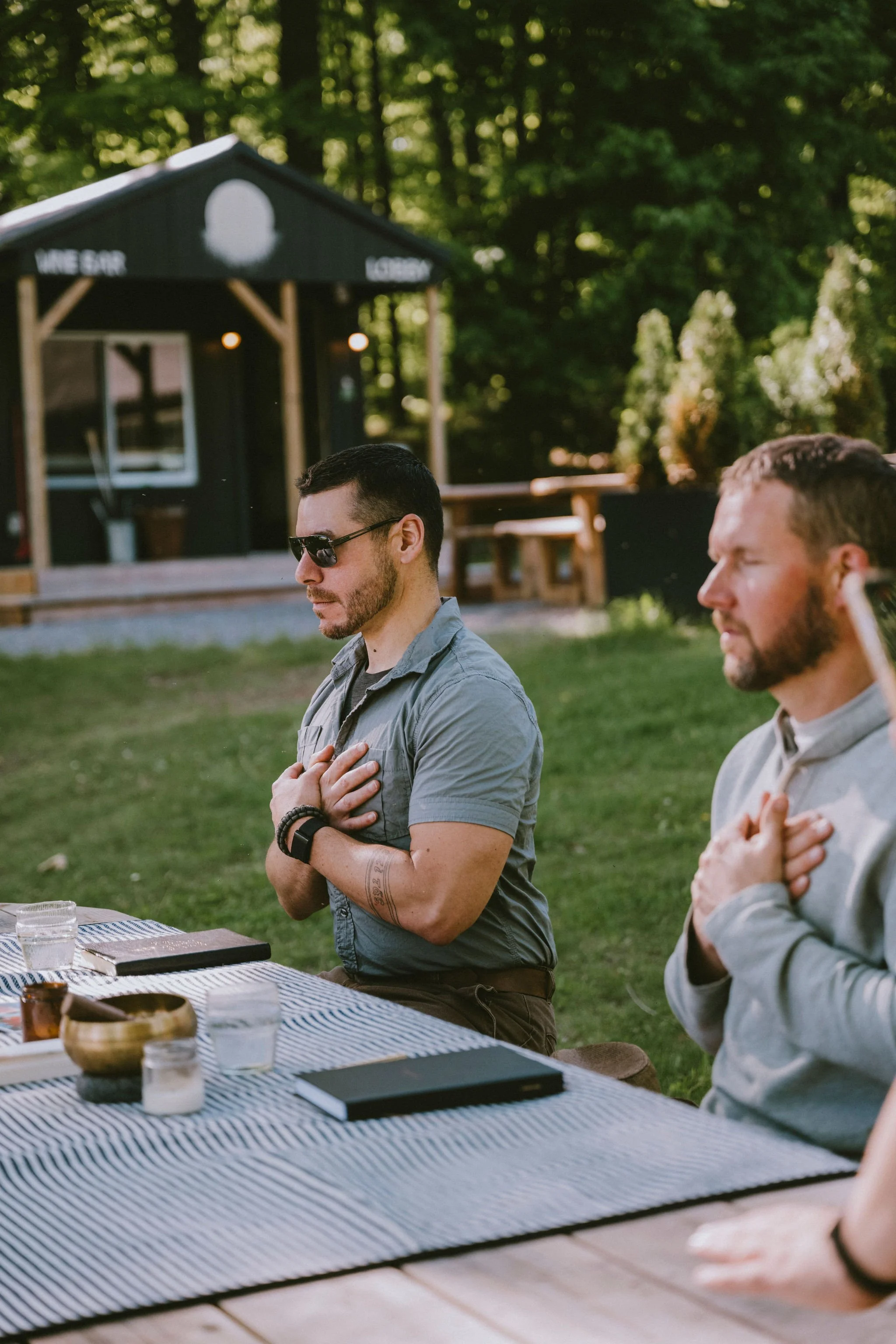 Two men sitting at a table outdoors with their hands over their hearts, praying or reflecting, near a black cabin in a wooded area.