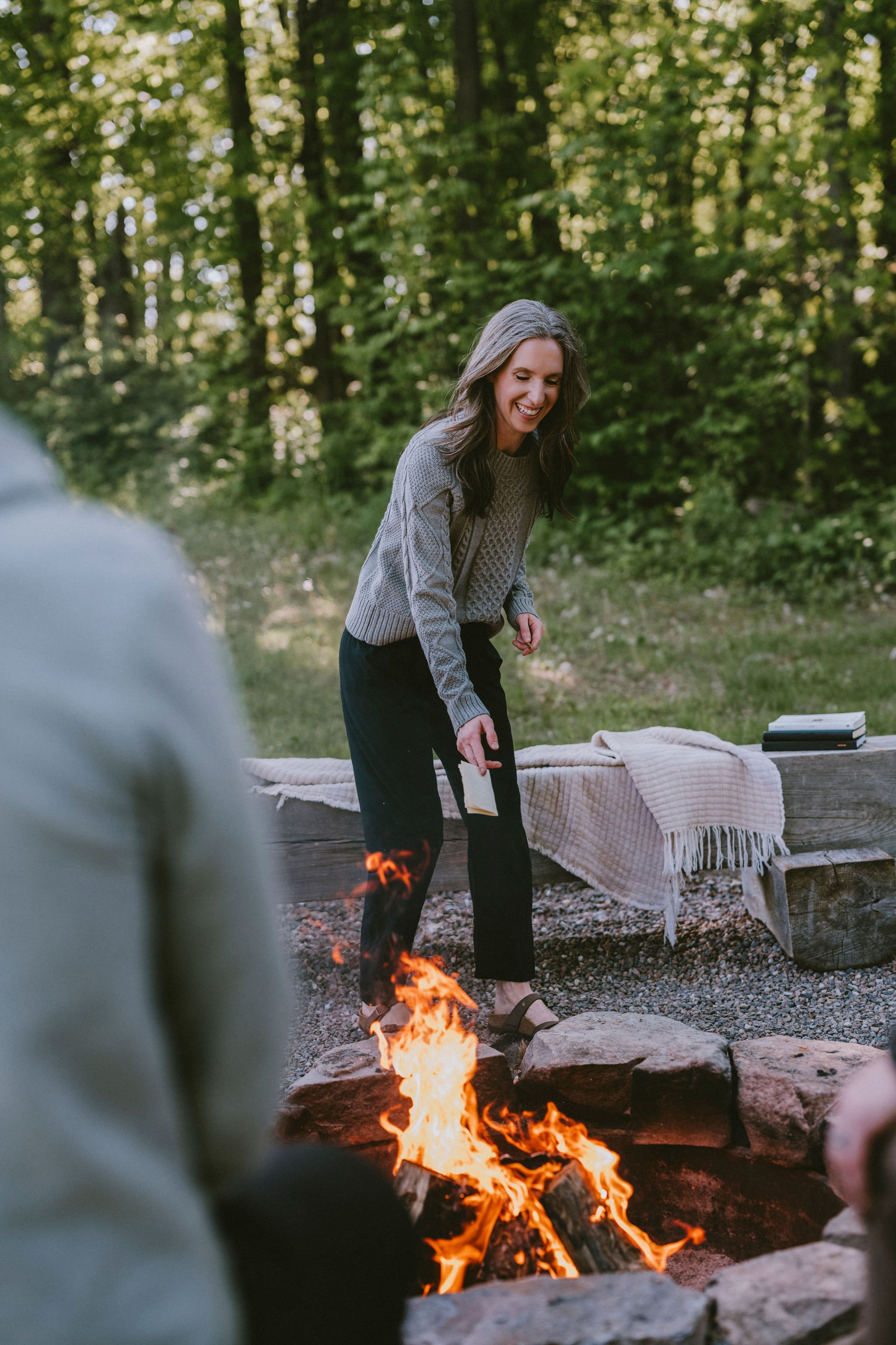 A woman smiling and leaning slightly forward, reaching towards a campfire, outdoors in a wooded area. She is wearing a gray sweater and black pants, with a plaid blanket on a wooden bench beside her.