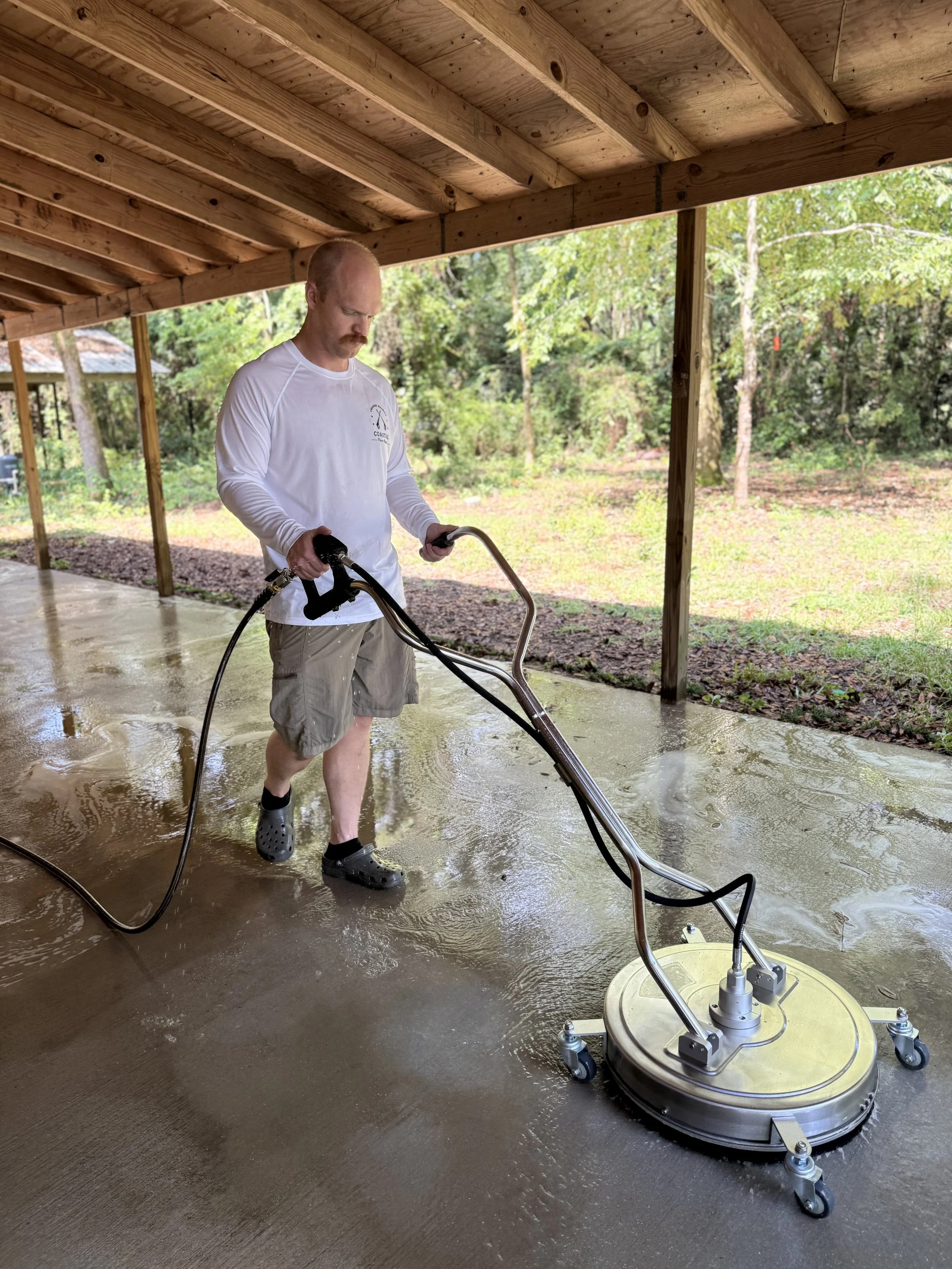 A man pressure washing a concrete patio under a wooden porch, with trees and grass in the background.