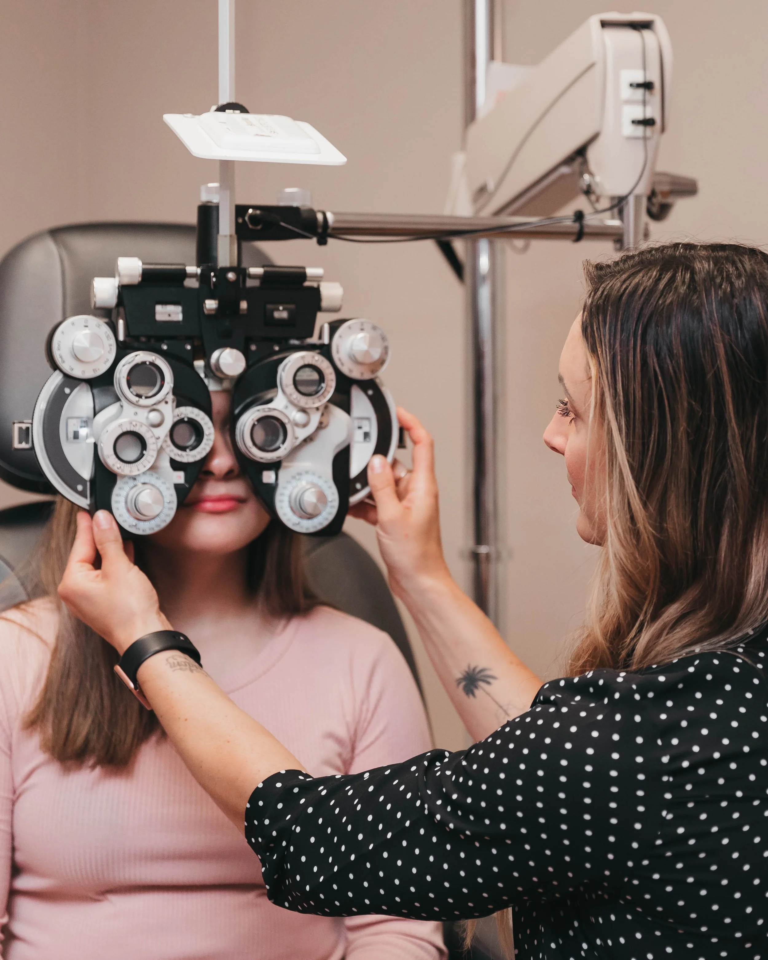 A woman at an eye appointment has her eyes checked with phoropter, while an eye care professional adjusts the equipment.