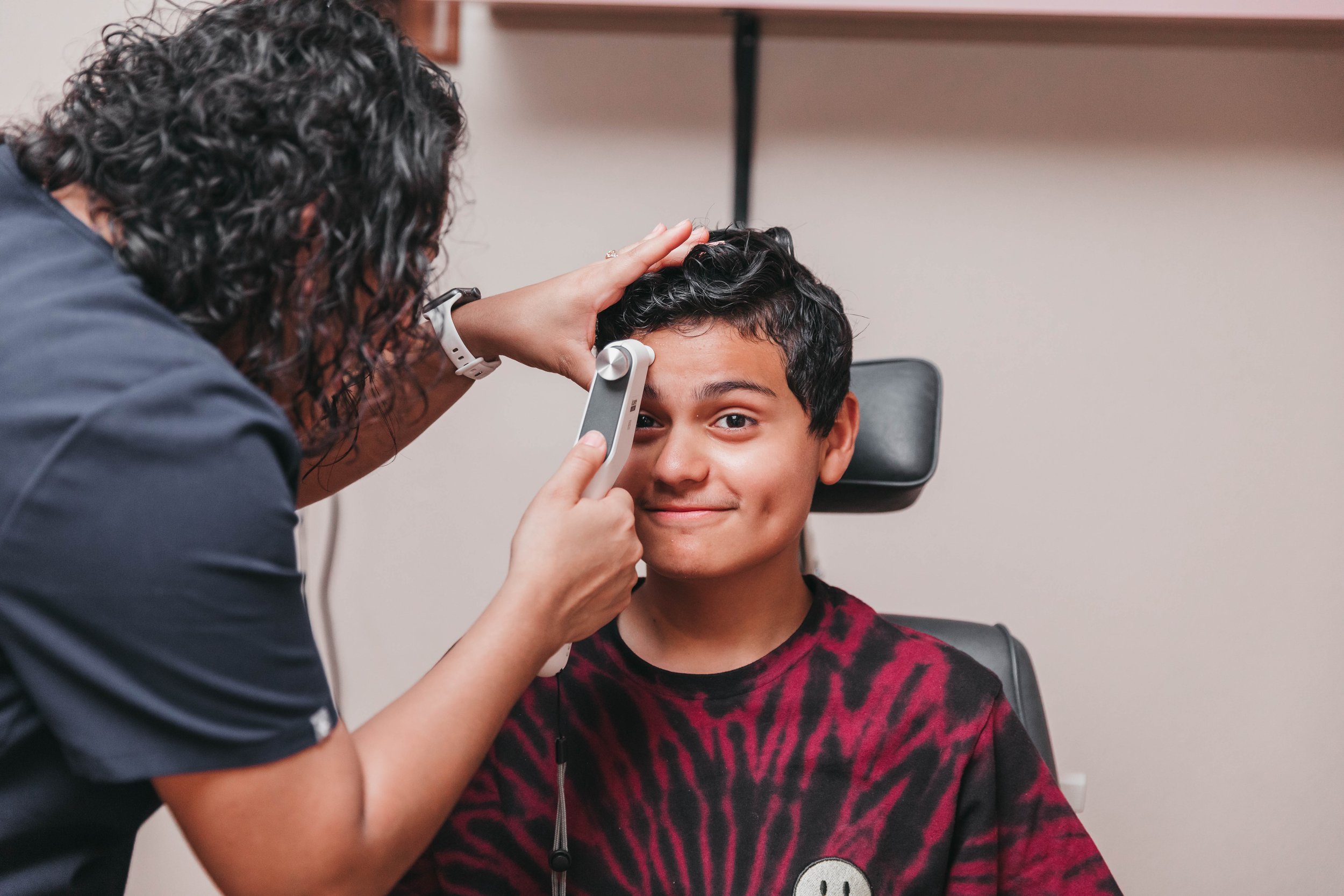 A healthcare professional examines a young boy's ear while he sits in a medical chair. The boy has short dark hair and is wearing a red and black patterned shirt. The healthcare worker has curly dark hair and is using an otoscope to look inside the boy's ear in a clinical setting.
