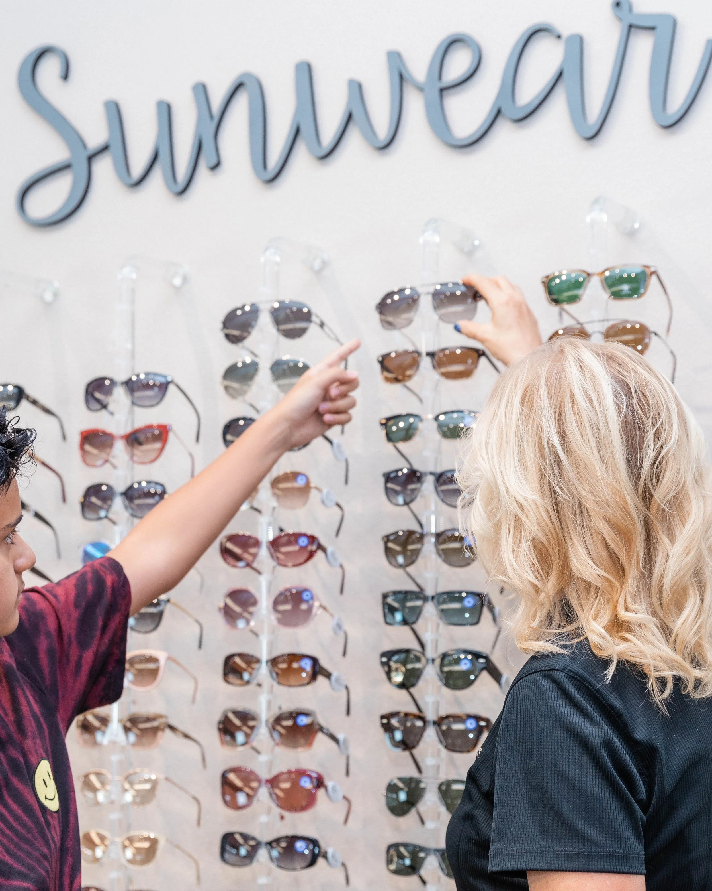 Two people shopping for sunglasses at a retail store, with a wall of sunglasses display and a large sign that says 'sunglasses' in the background.