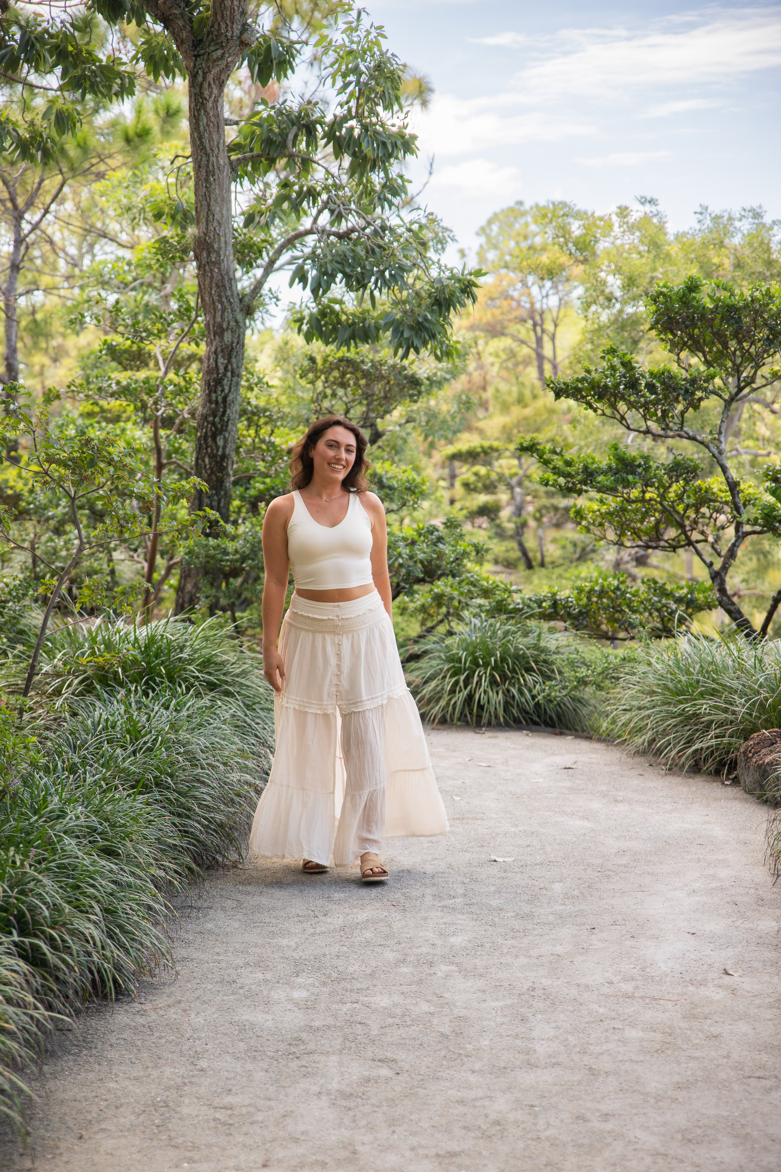 A woman dressed in white, walking on a garden pathway lined with green bushes and trees, smiling at the camera.