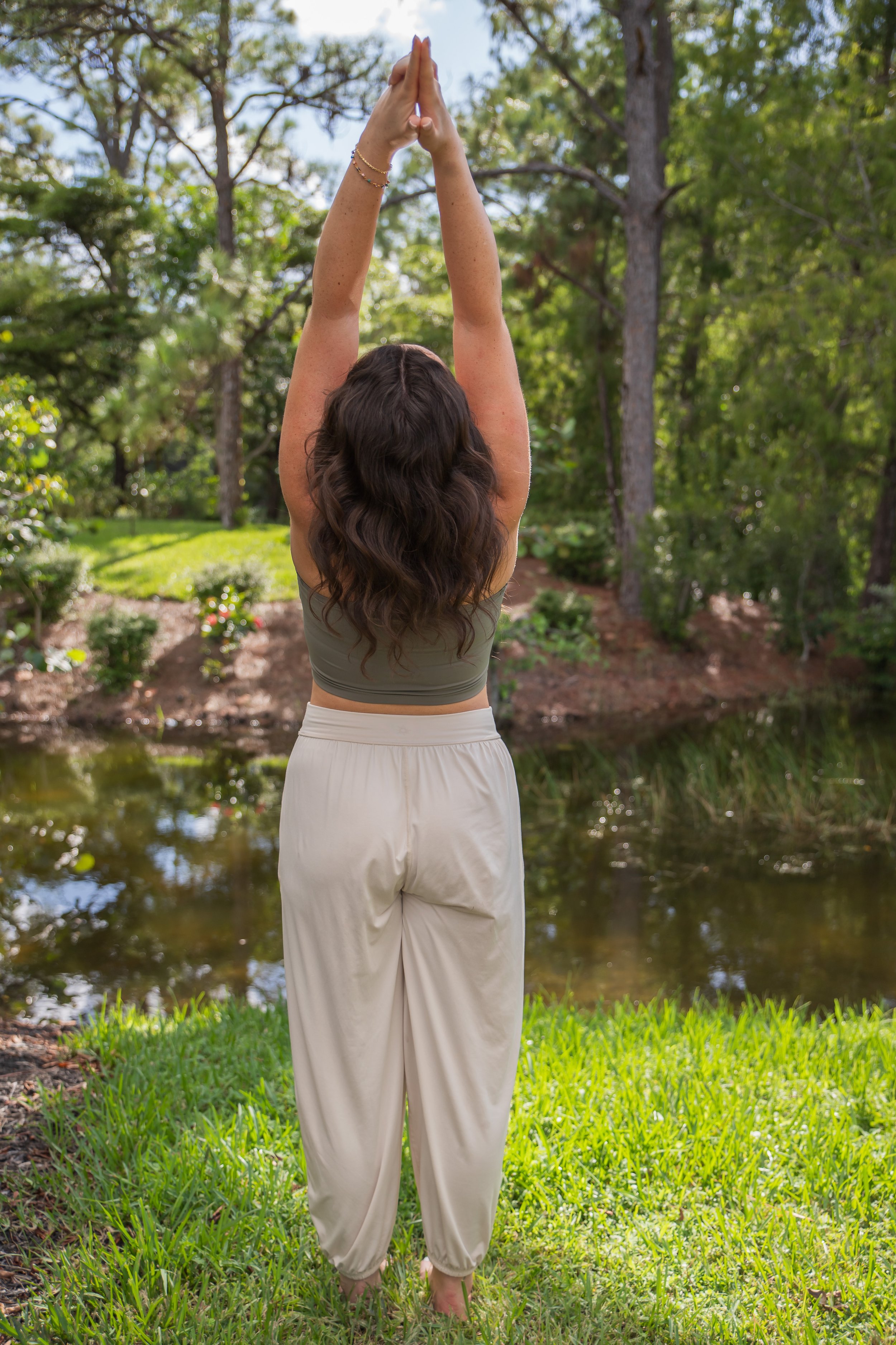 A woman standing on grass near a pond, practicing yoga with her arms extended upward and palms together, surrounded by trees and greenery.