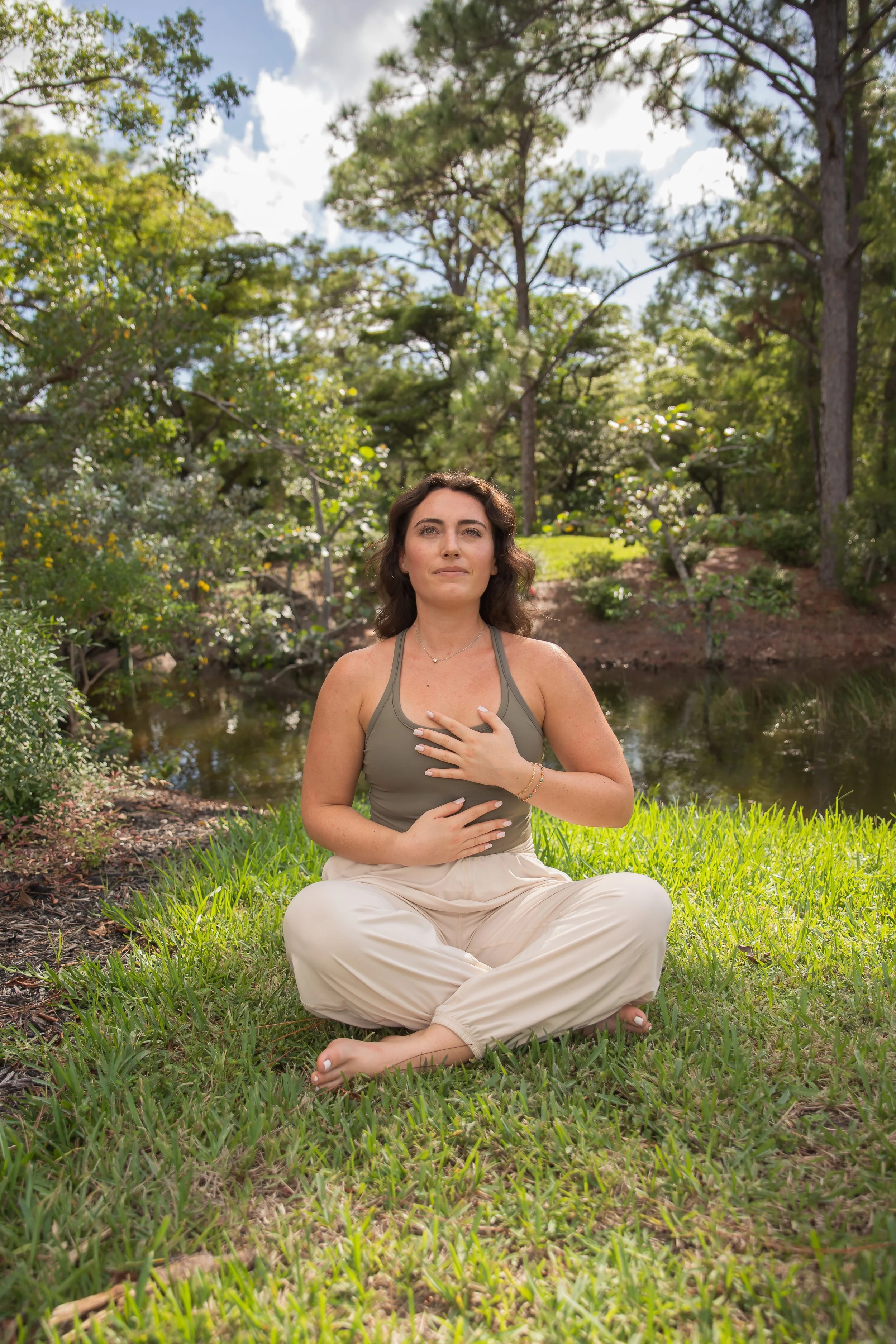 A woman practicing meditation outdoors on a grassy area near a pond, surrounded by trees and greenery, during daytime.