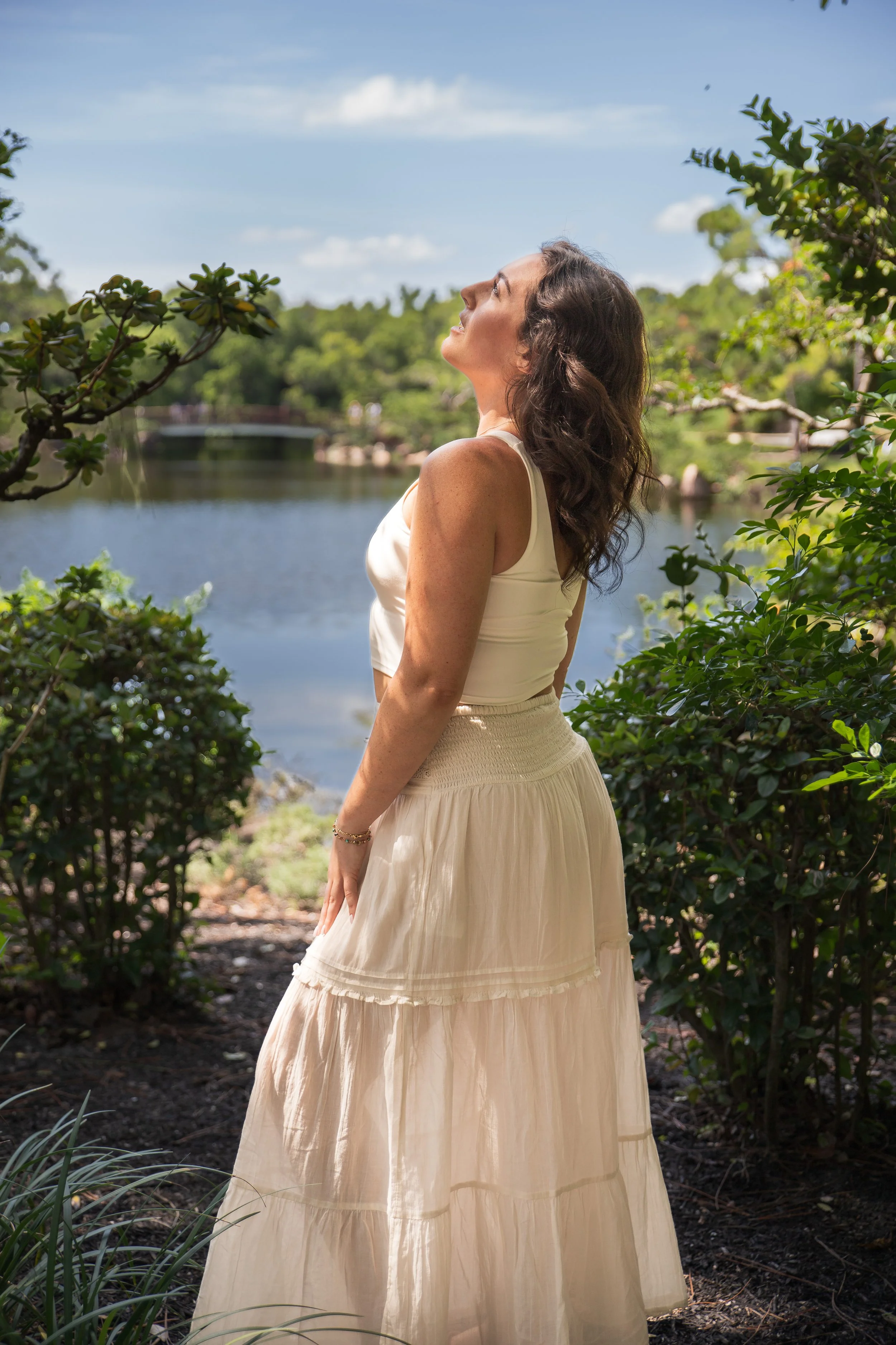 A woman in a white dress standing outdoors near water, with her head tilted back and eyes closed, surrounded by greenery.
